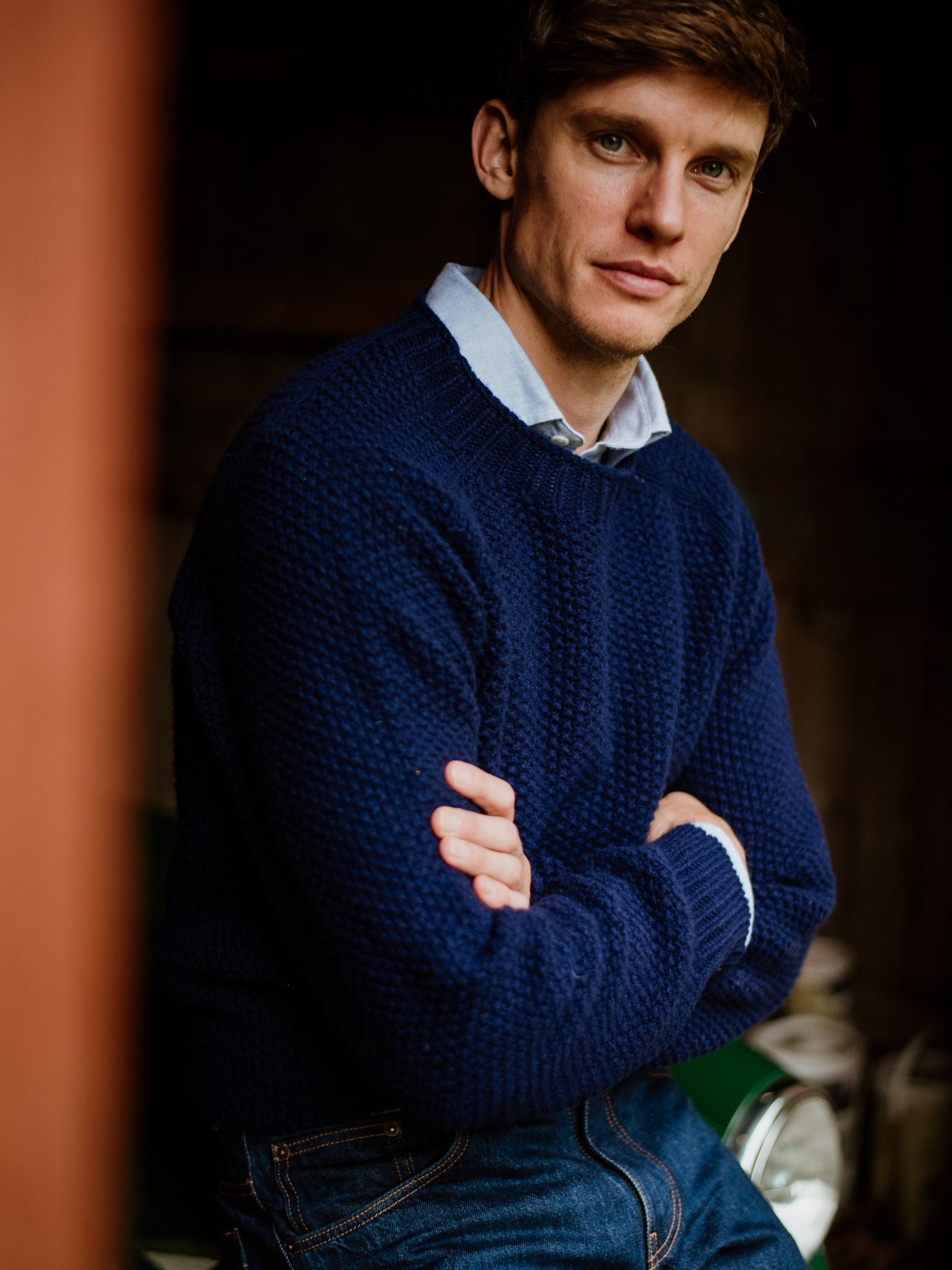 A man with light brown hair wears a Campbell's of Beauly Moss Stitch Jumper over a collared shirt and jeans. He sits with arms crossed, looking calmly at the camera.