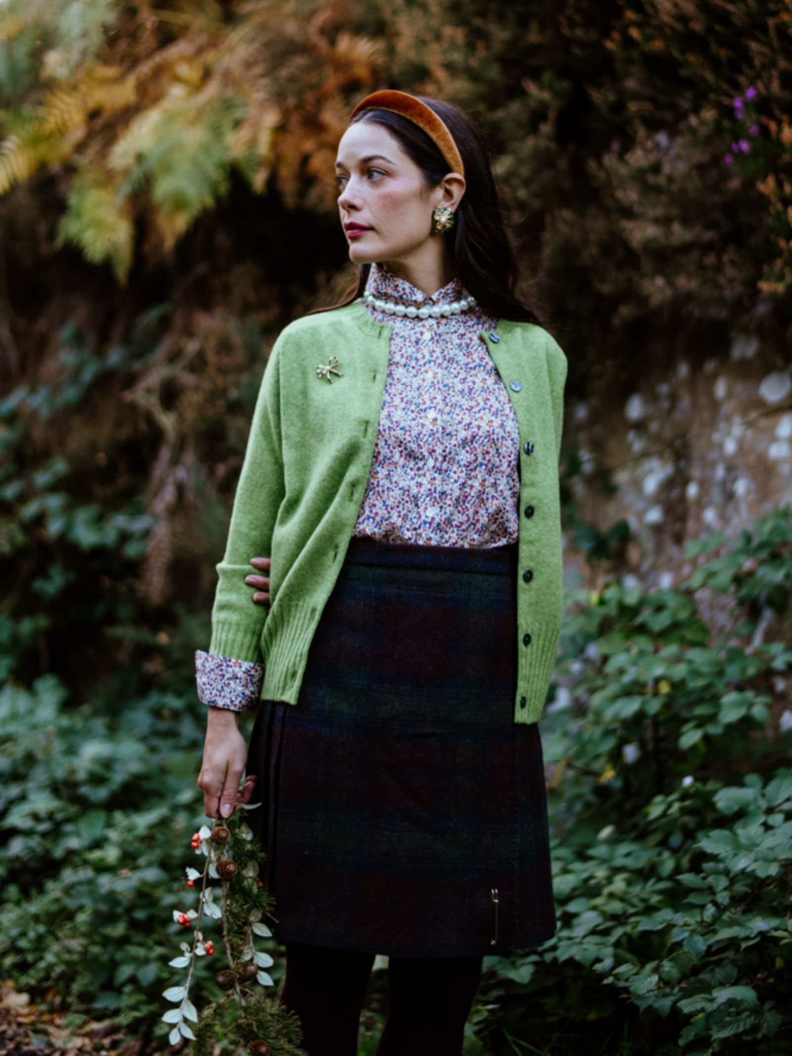 A woman stands outside by greenery, wearing the Campbell's of Beauly Geelong Lambswool Crew Cardigan over a floral blouse with a dark skirt and brown headband. She holds a berry branch and gazes thoughtfully to the side.