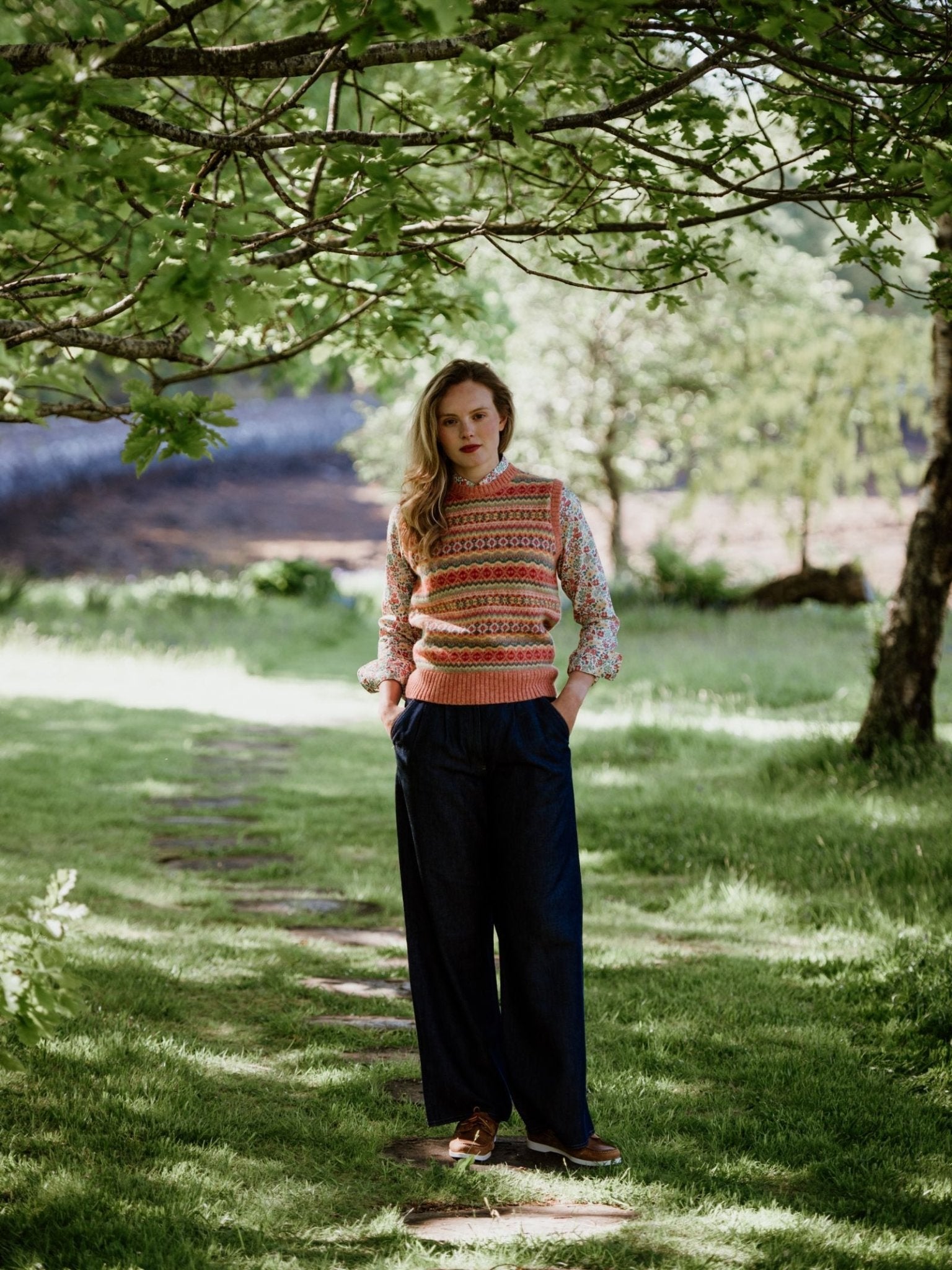 A woman stands on a stone path in a sunlit garden, wearing Campbells of Beauly Wide Leg Jeans with a patterned knit vest and floral shirt, hands in her pockets amid green trees and grass.