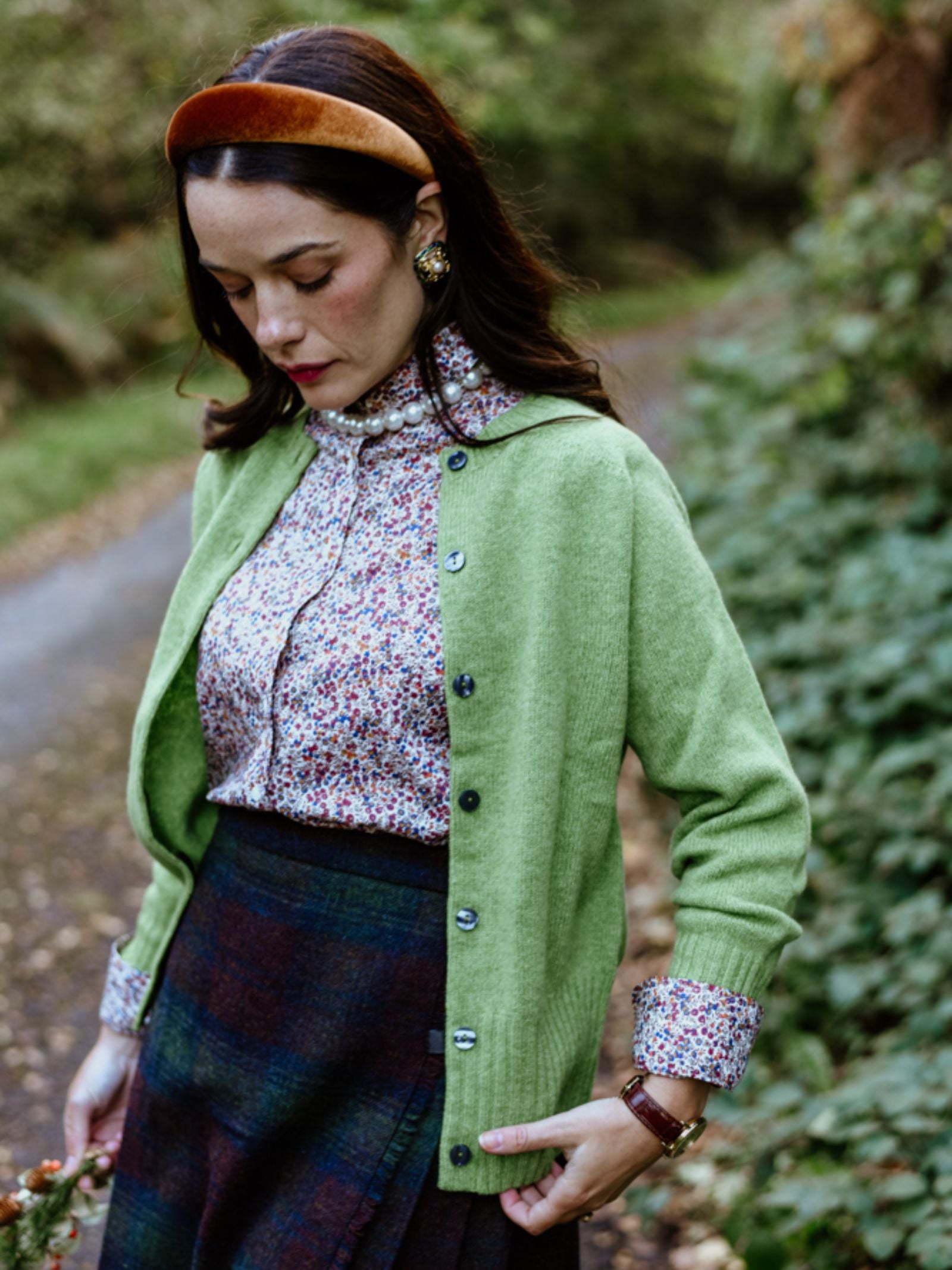 A woman with dark hair wears a green Campbell's of Beauly Geelong Lambswool Crew Cardigan over a floral blouse and plaid skirt, standing on a path amid greenery, looking down with her hands in her pockets.