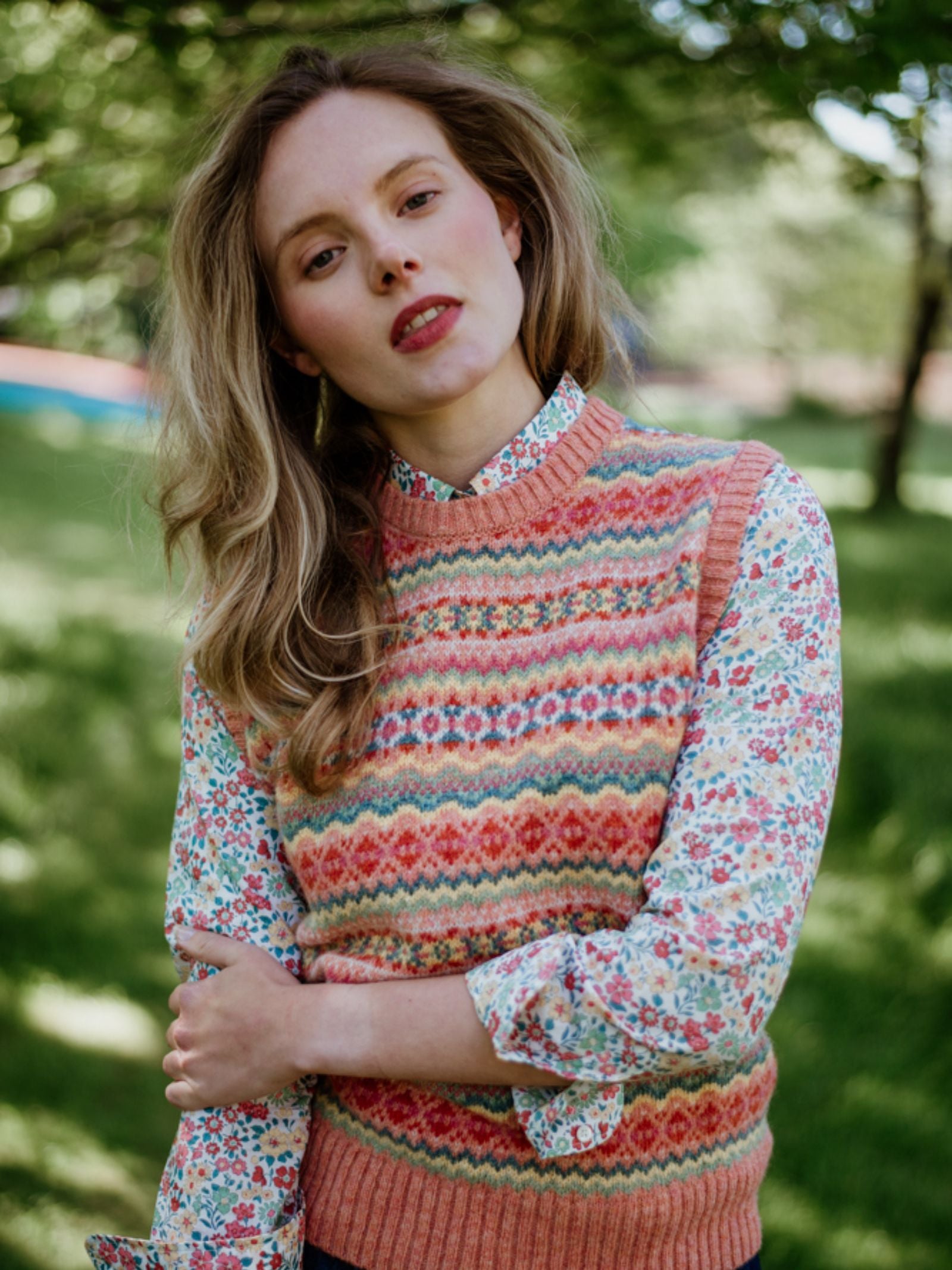 A woman with long blonde hair stands outdoors among sunlit greenery, wearing a Campbells of Beauly Fairisle Crew Tank in vibrant Shetland wool over a floral shirt, her expression relaxed.