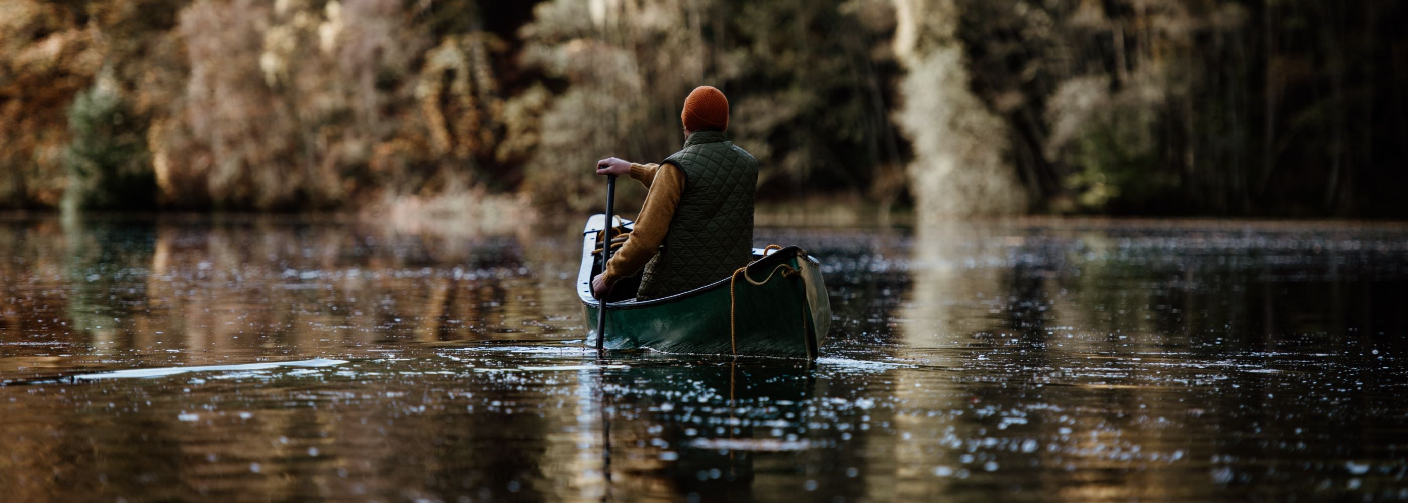 A person wearing a beanie and vest paddles a canoe on a calm lake surrounded by autumn trees, with leaves floating on the water.