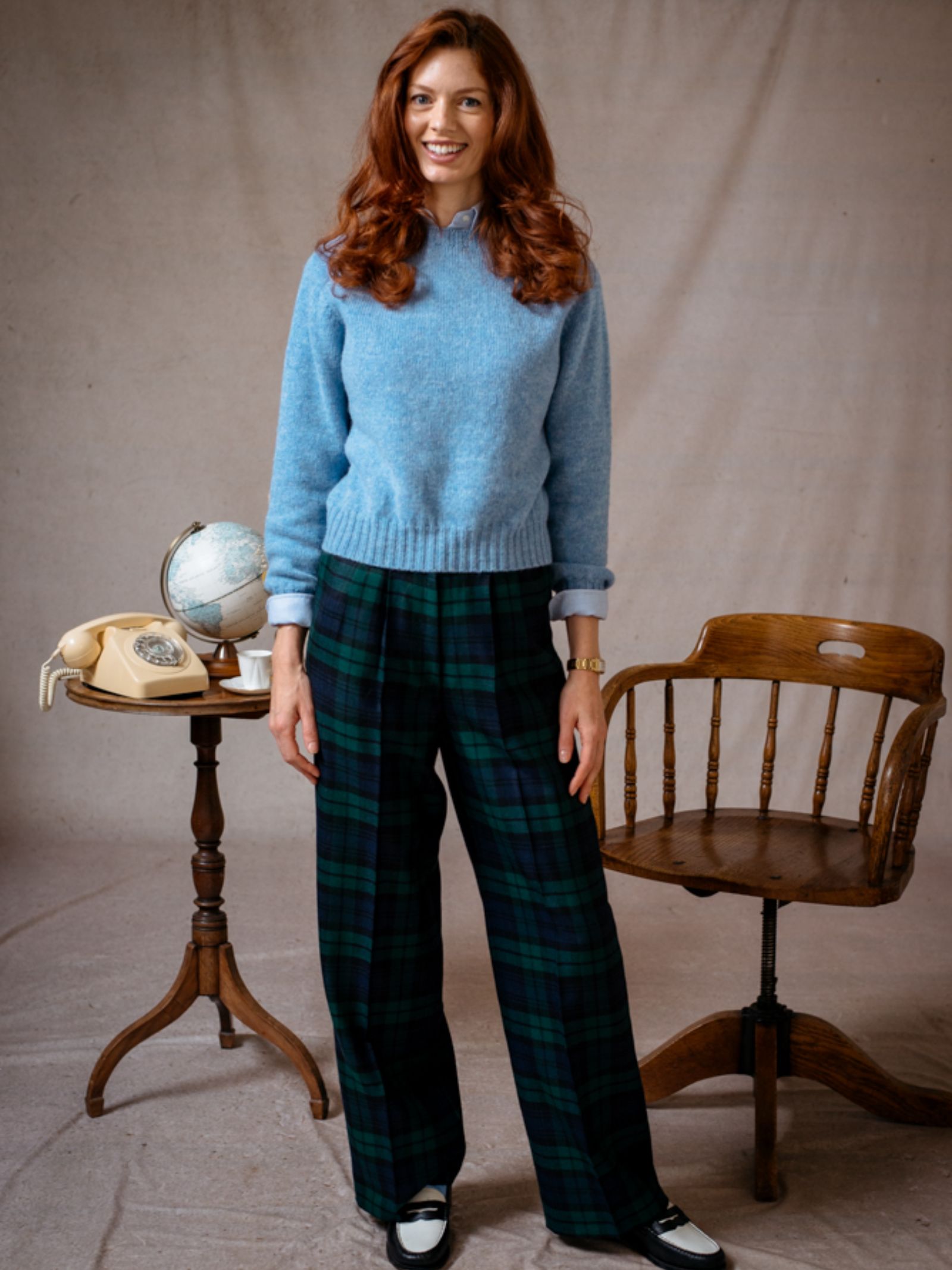 A woman with long red hair smiles indoors, wearing a light blue Cropped Shetland Jumper by Campbells of Beauly and green plaid pants, standing beside a wooden chair and a small table with a rotary phone and globe.