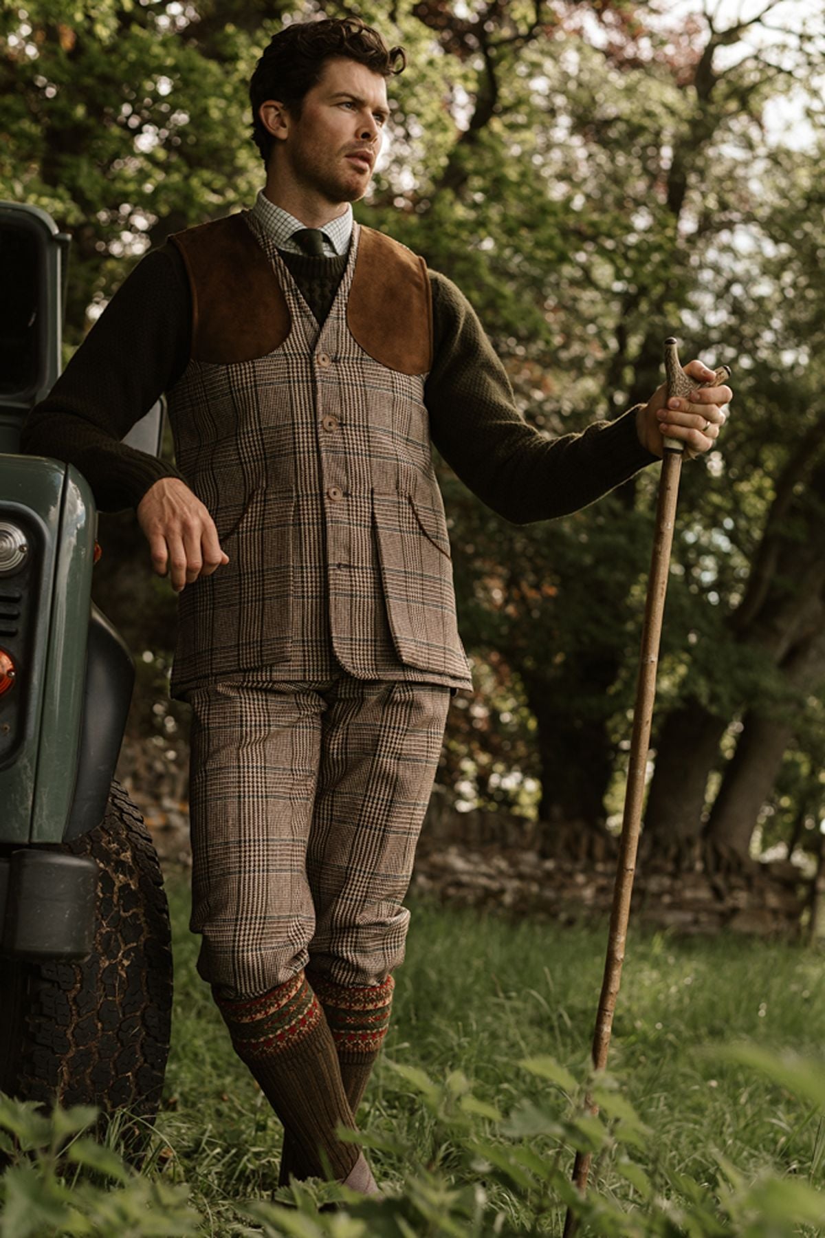Man in a tweed waistcoat and trousers, green jumper, and patterned knee-high socks leaning on a walking stick beside a vintage car.