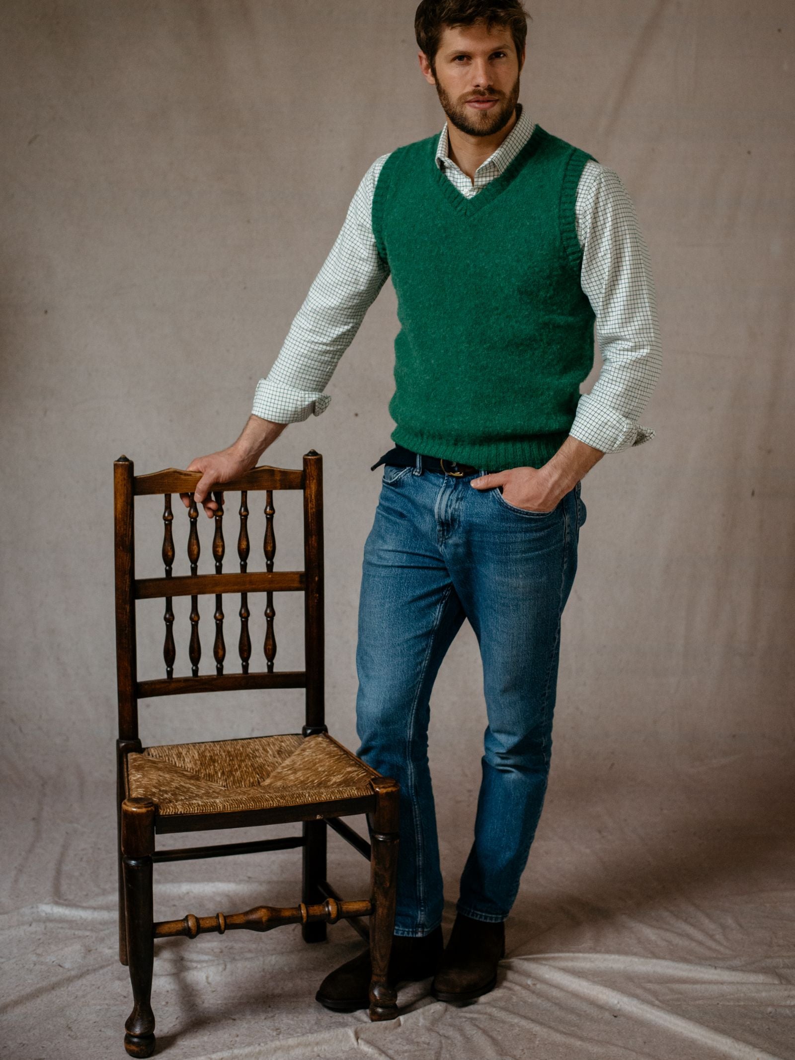 A man wearing the Campbells of Beauly Shetland Slipover stands by a wooden chair with a woven seat, resting one hand on the chair back, against a plain fabric backdrop.