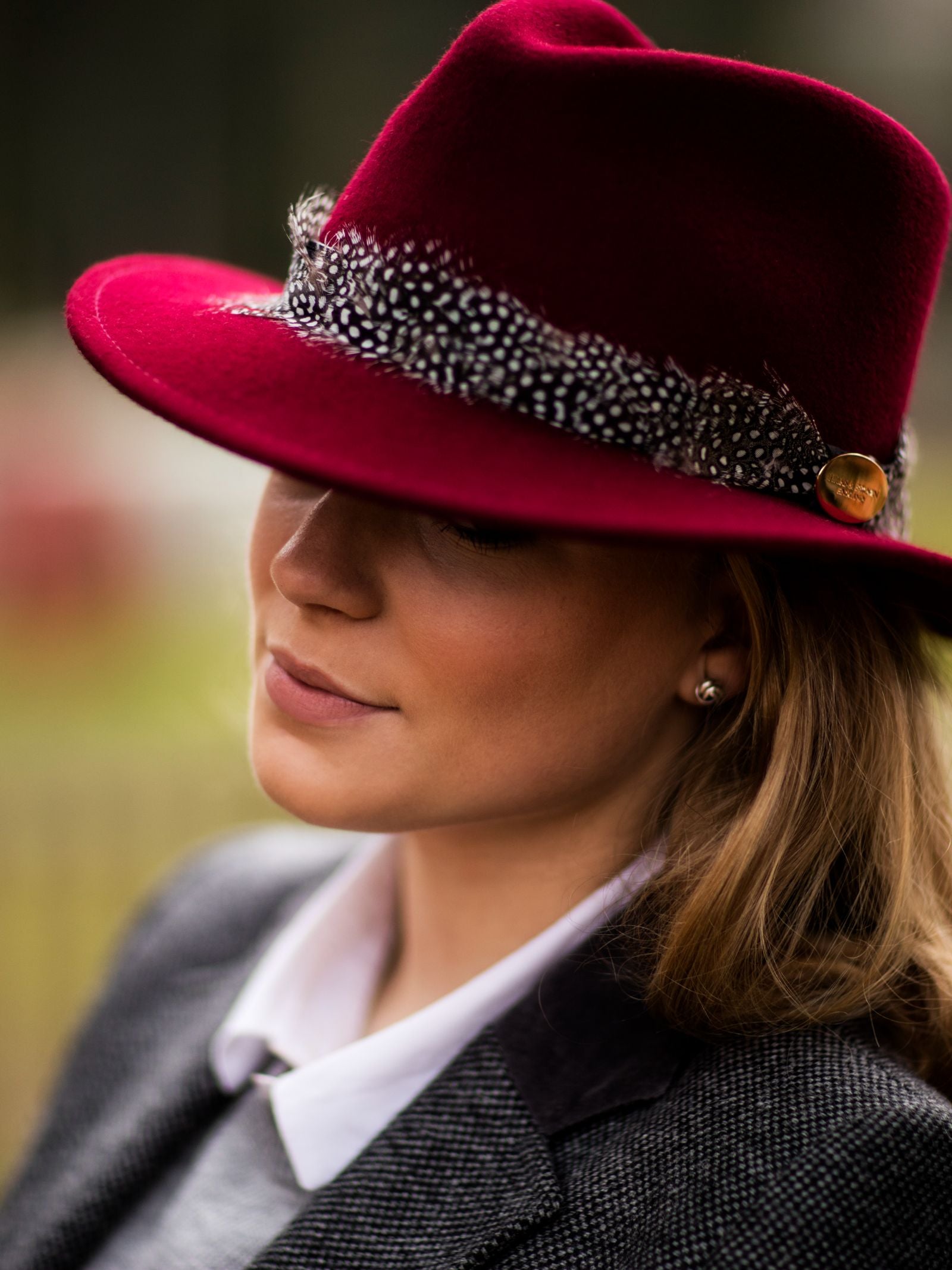 A woman wears the Hicks & Brown Guinea Feather Maroon Fedora with a gold pin, styled with a gray blazer and white collared shirt. She gazes down softly, framed by a gently blurred background.