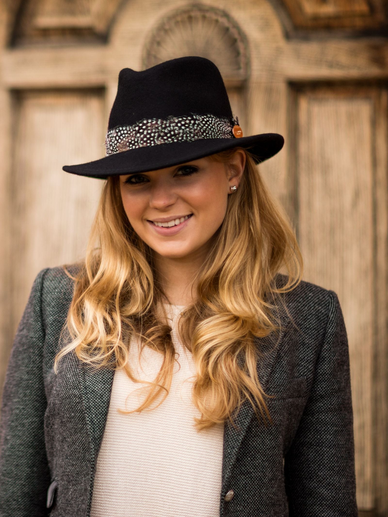 A smiling woman with long blonde hair wears the Hicks & Brown Guinea Feather Maroon Fedora, paired with a grey blazer and light sweater, as she stands in front of a carved wooden door.