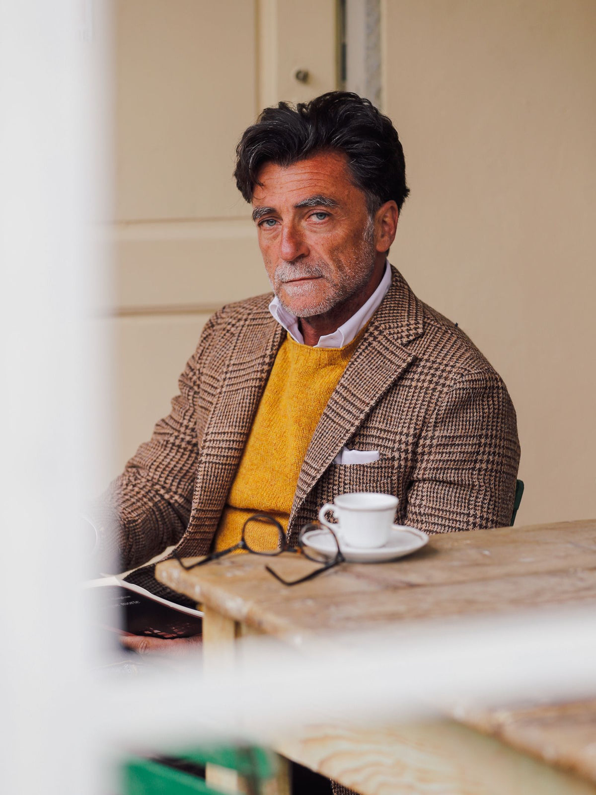 A man with dark hair and a beard, dressed in a Campbell’s of Beauly Harris Tweed Jacket in Glencheck, featuring full canvas construction and paired with a yellow sweater, sits at a wooden table with a cup and glasses, looking thoughtfully at the camera.