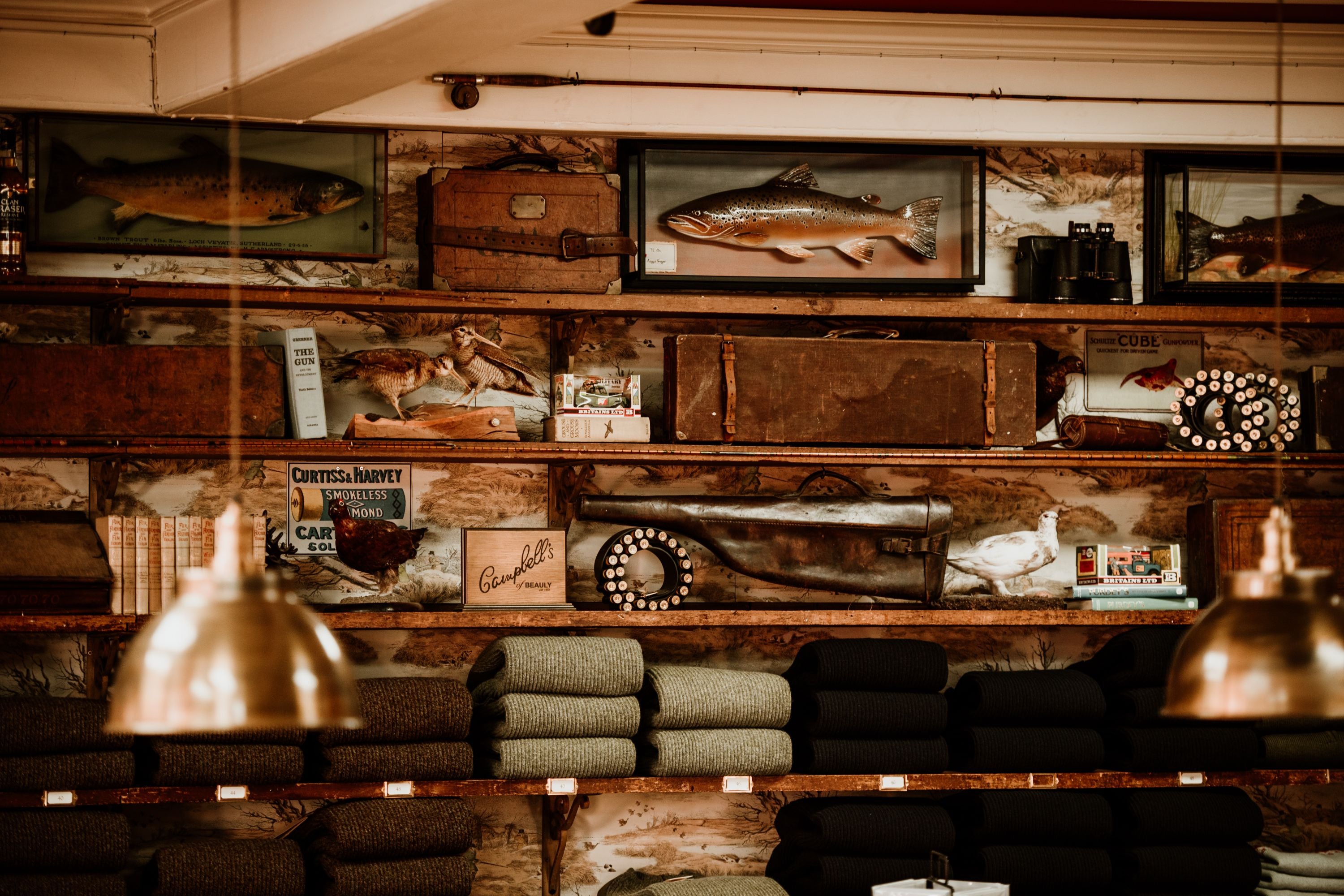 A cozy shop interior features shelves lined with folded woolen blankets, vintage suitcases, books, fishing reels, old cameras, and mounted fish, with warm light from hanging lamps.