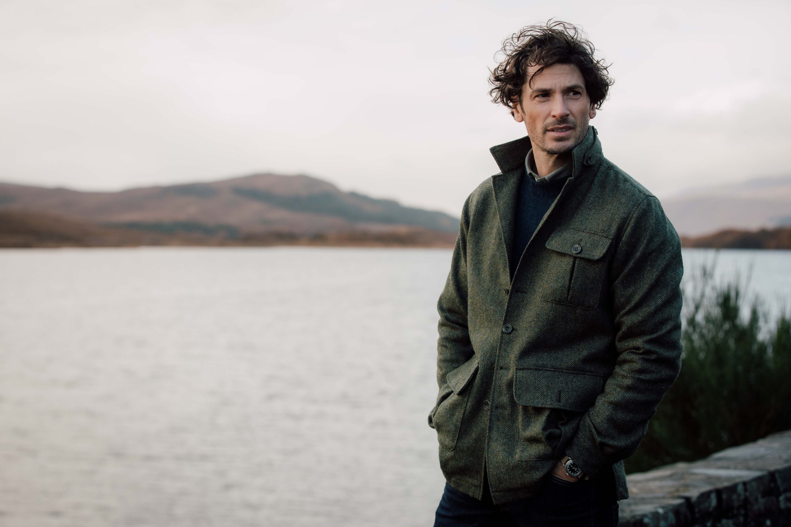 A man with curly hair wearing a green jacket stands by a lake with mountains in the background. He has his hands in his pockets and looks off to the side, with overcast skies above.