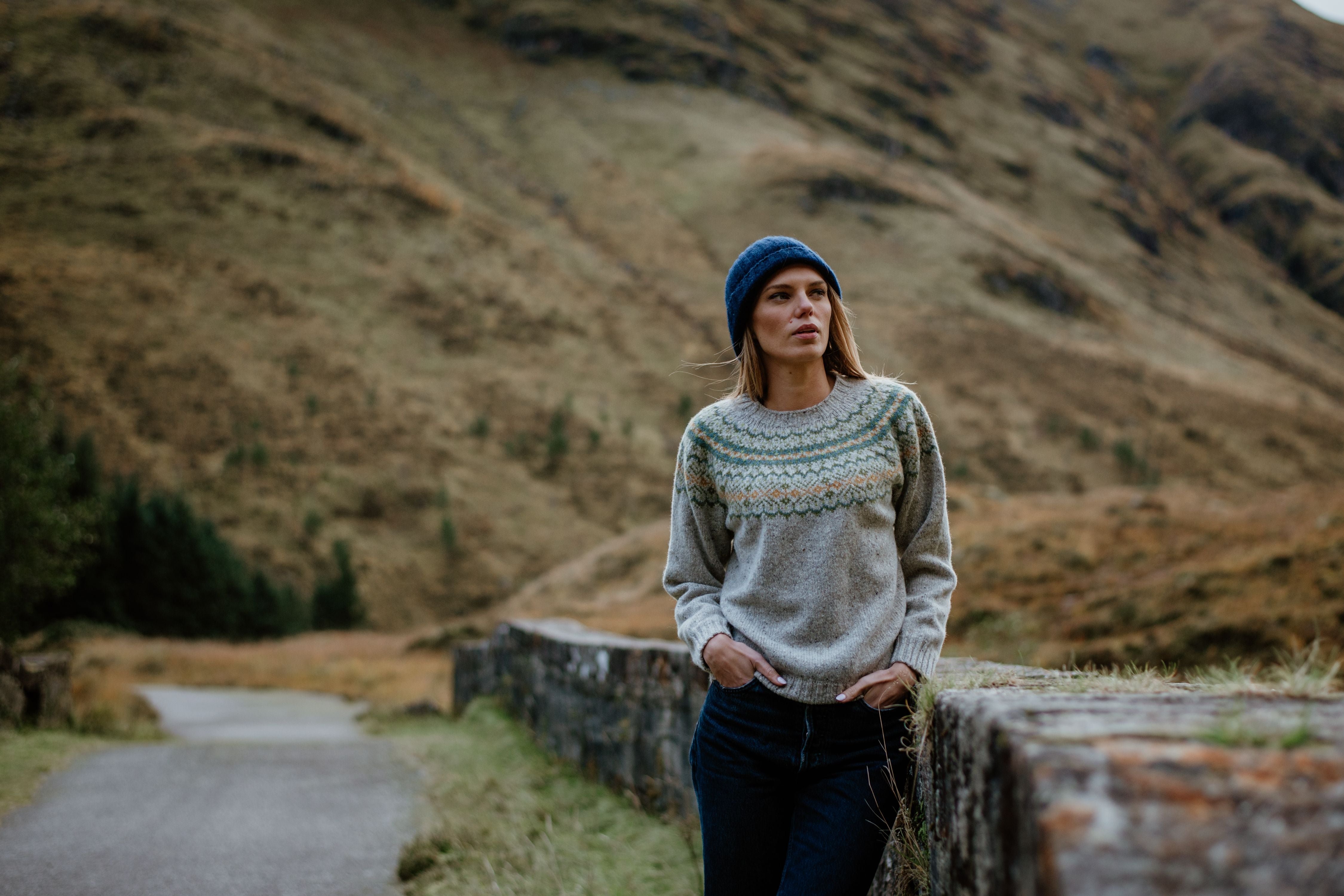 A woman wearing a knitted sweater and blue beanie stands outdoors, leaning on a stone wall with a mountainous, grassy landscape in the background.