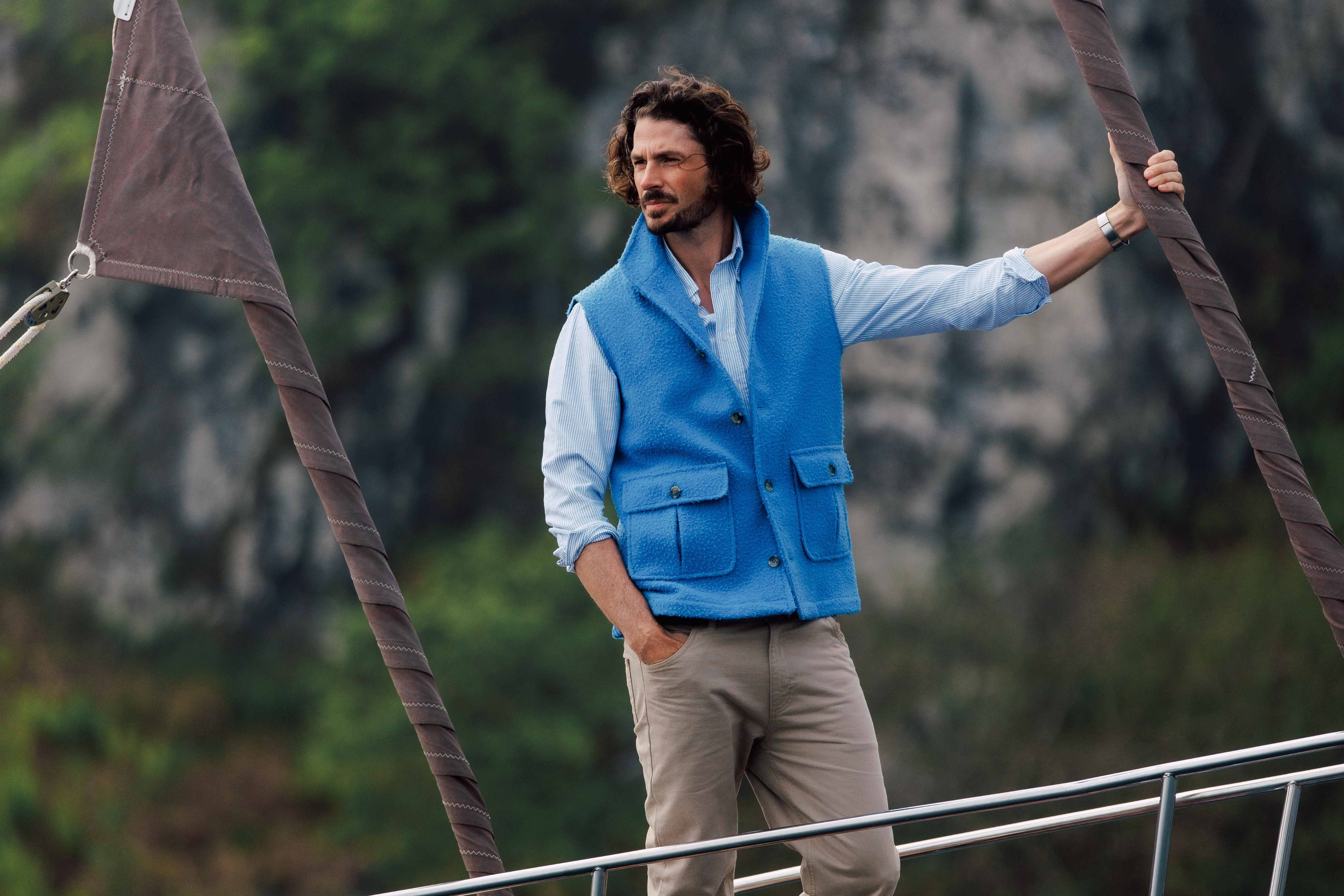 A man with wavy hair, wearing a light blue shirt and bright blue fleece vest, stands on a boat outdoors, holding onto ropes, with a blurred natural background of trees and rocks.