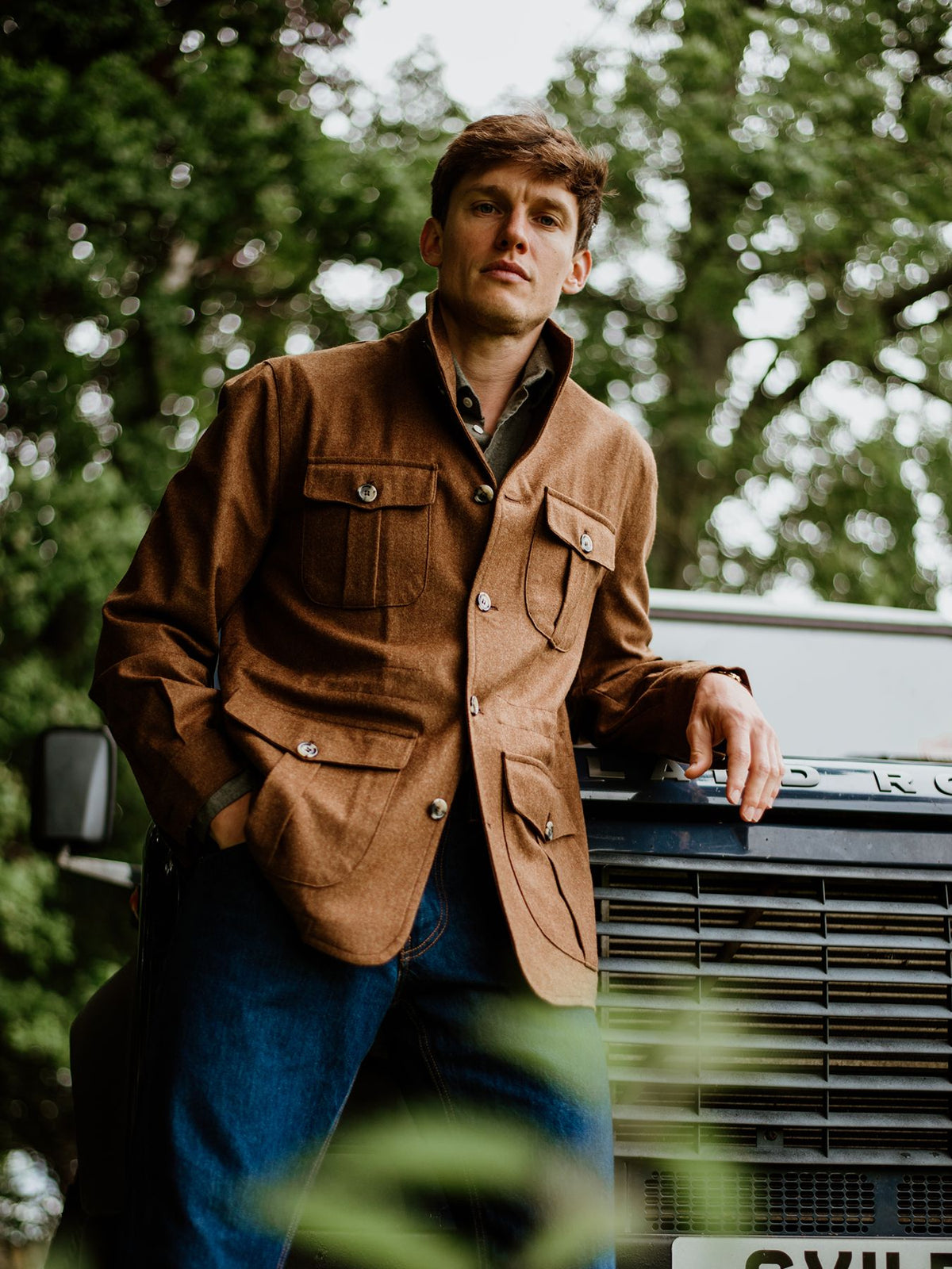 A man wearing a brown four-pocket field jacket and blue jeans, leaning against the front of a dark vehicle.
