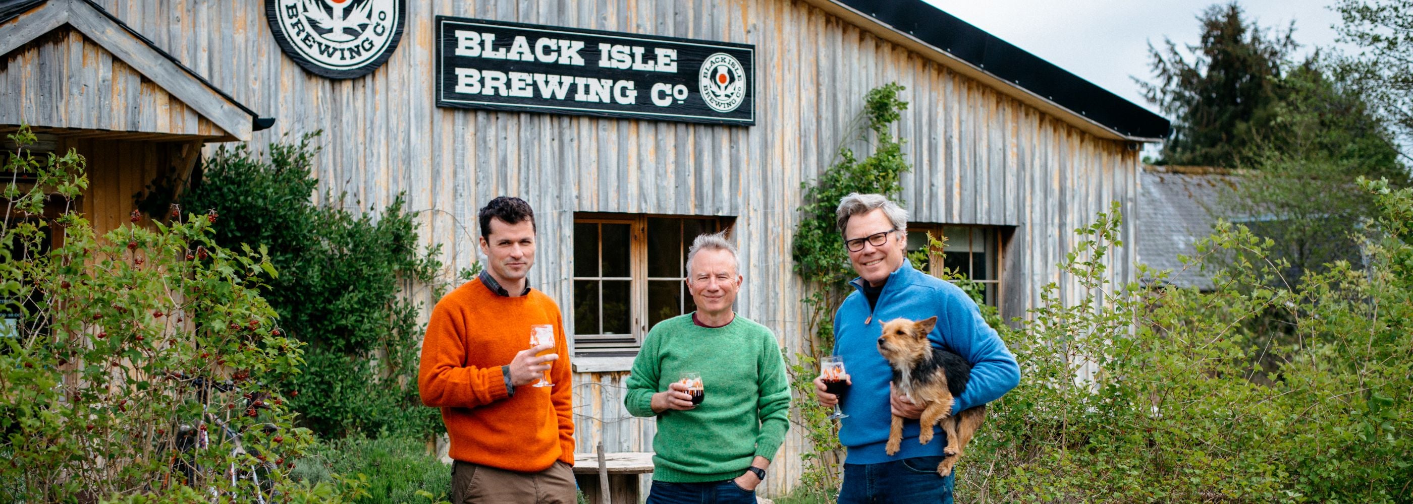 Three men stand outside a rustic wooden building with a “Black Isle Brewing Co” sign, each holding a drink. One man holds a small dog. Trees and greenery surround the scene.
