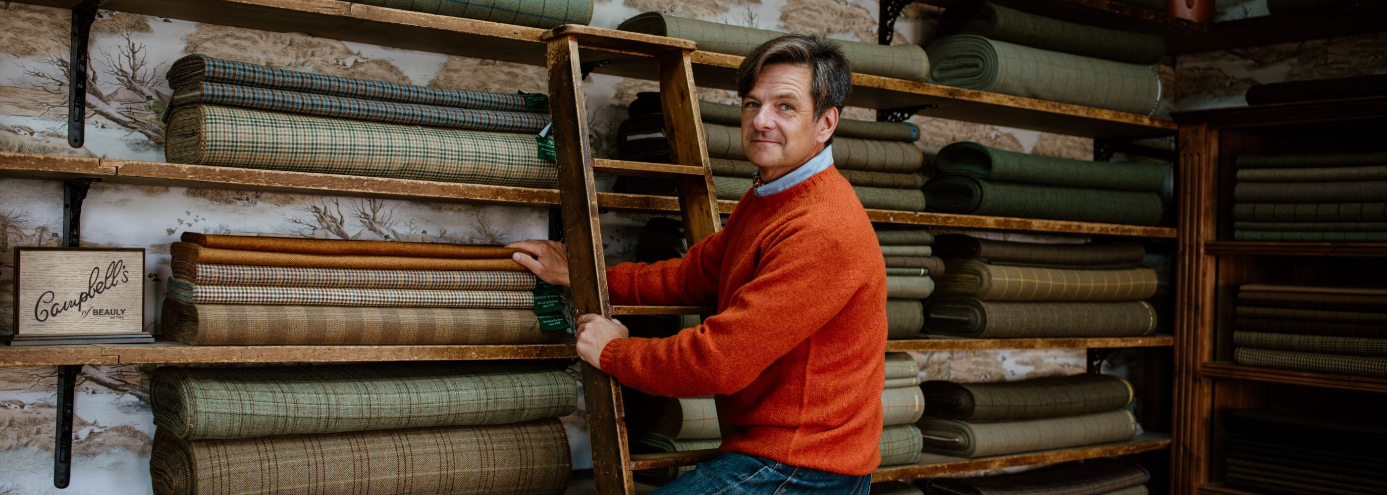 A man in an orange sweater sits on a wooden ladder in front of shelves filled with neatly stacked rolls of plaid and solid-colored fabric in various shades of green and brown.
