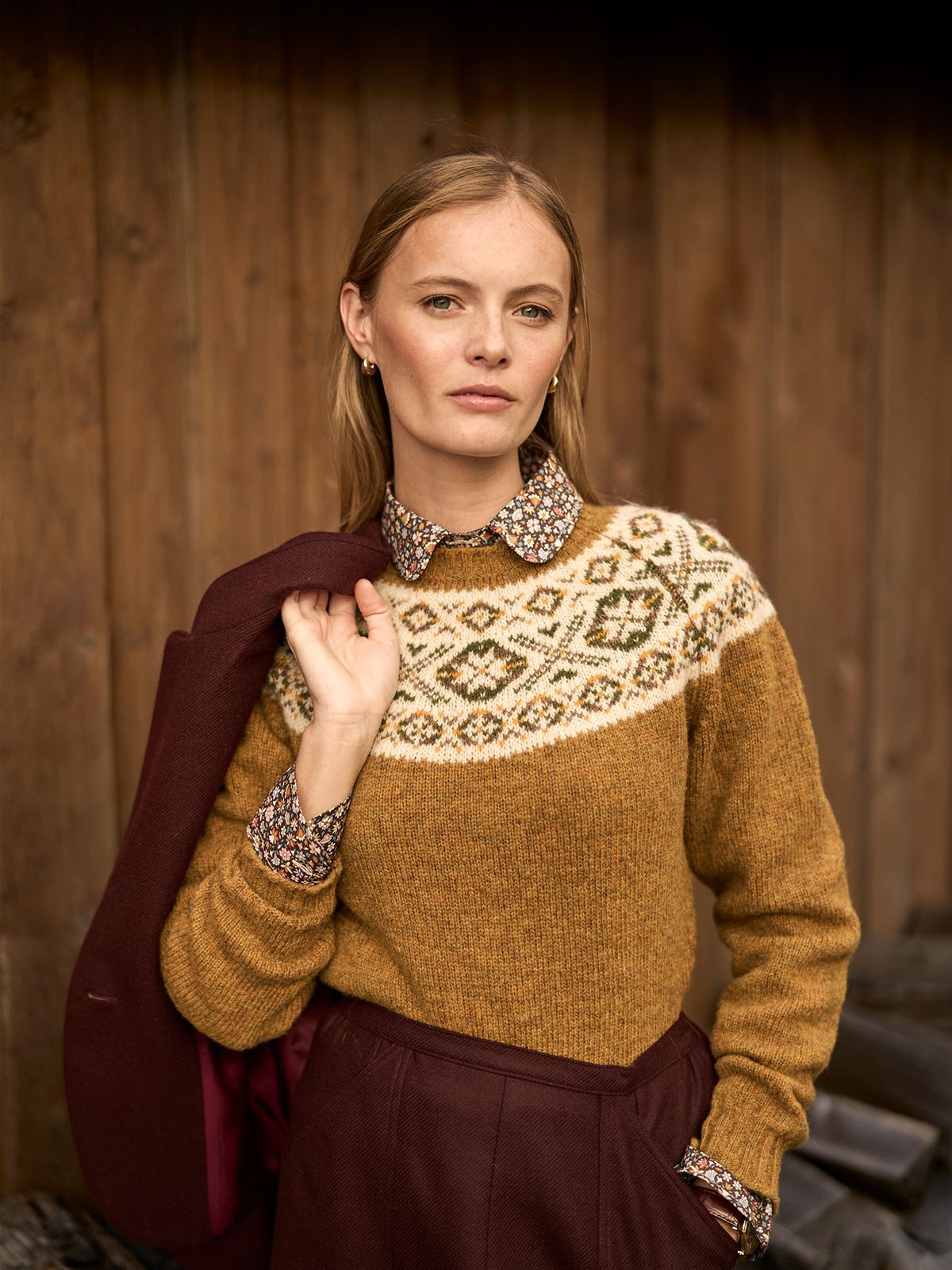A woman wearing a mustard-coloured Fair Isle jumper over a floral collared shirt, holding a maroon blazer over her shoulder.