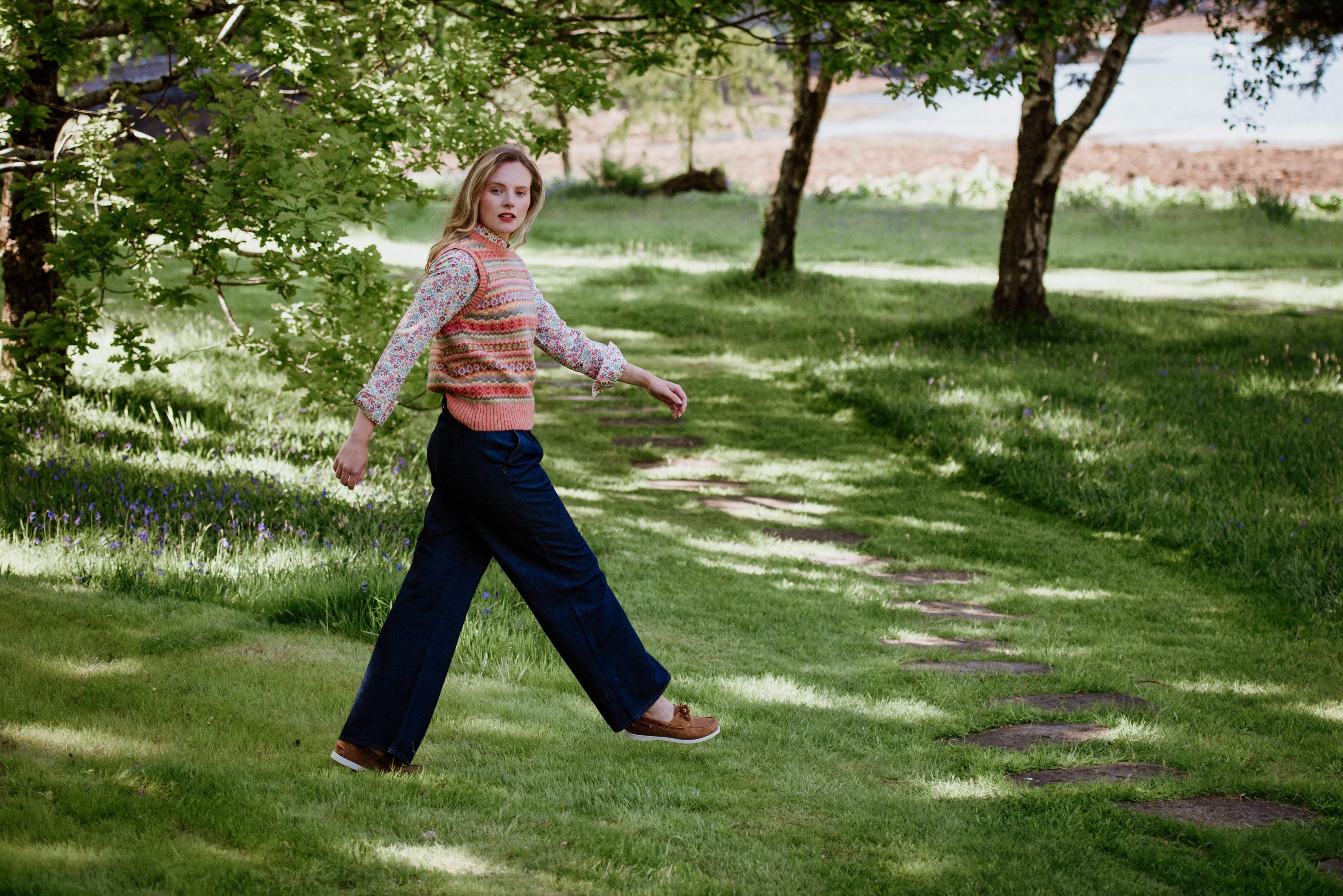 A woman with blonde hair, wearing a patterned sweater vest and wide-leg jeans, walks along a stone path in a grassy, tree-filled park, looking back over her shoulder.