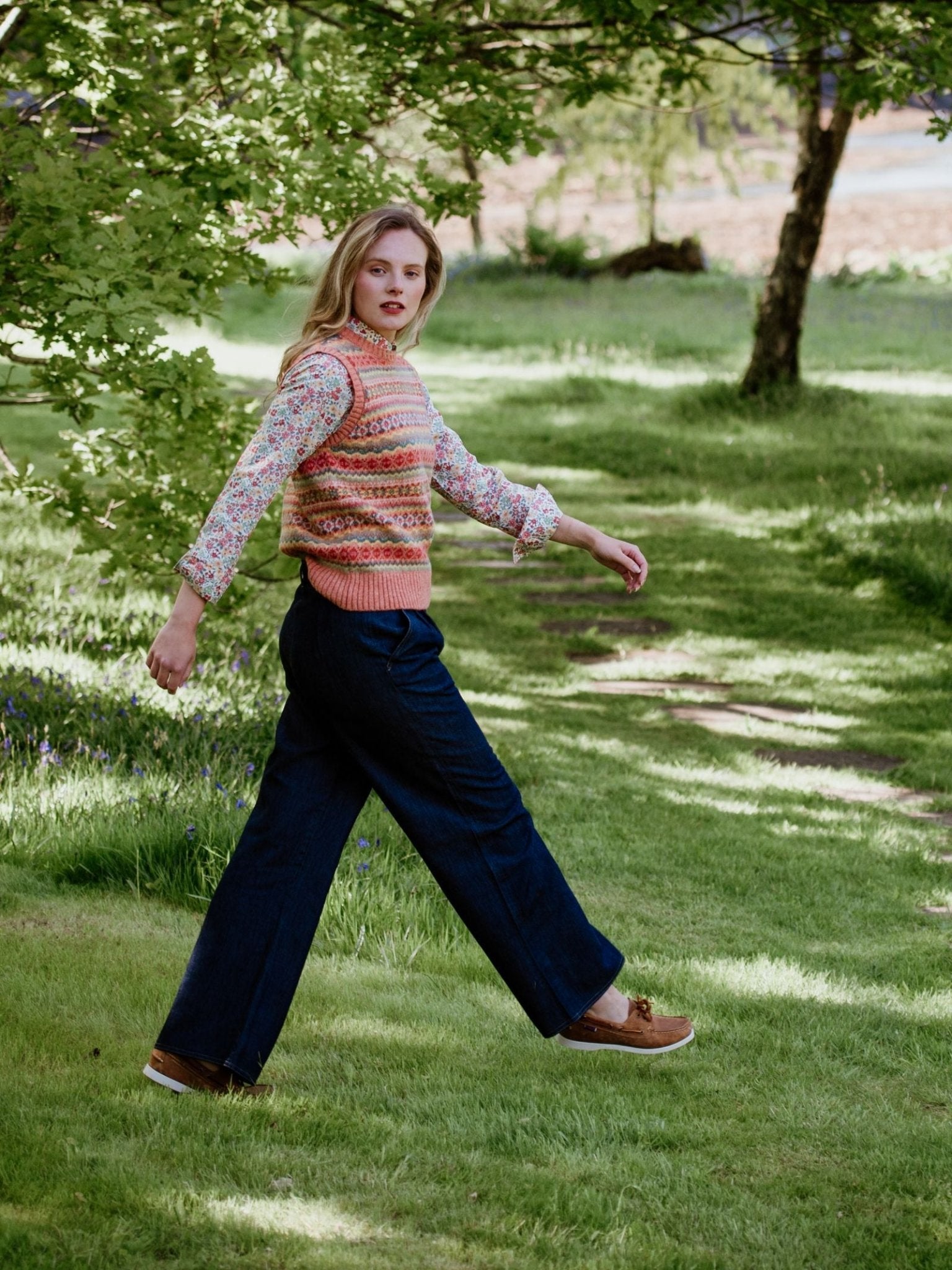 A woman with long blond hair walks on a grassy path, wearing Campbells of Beauly Wide Leg Jeans in lightweight denim, a patterned sleeveless sweater over a floral shirt, and brown shoes, with trees and sunlight in the background.