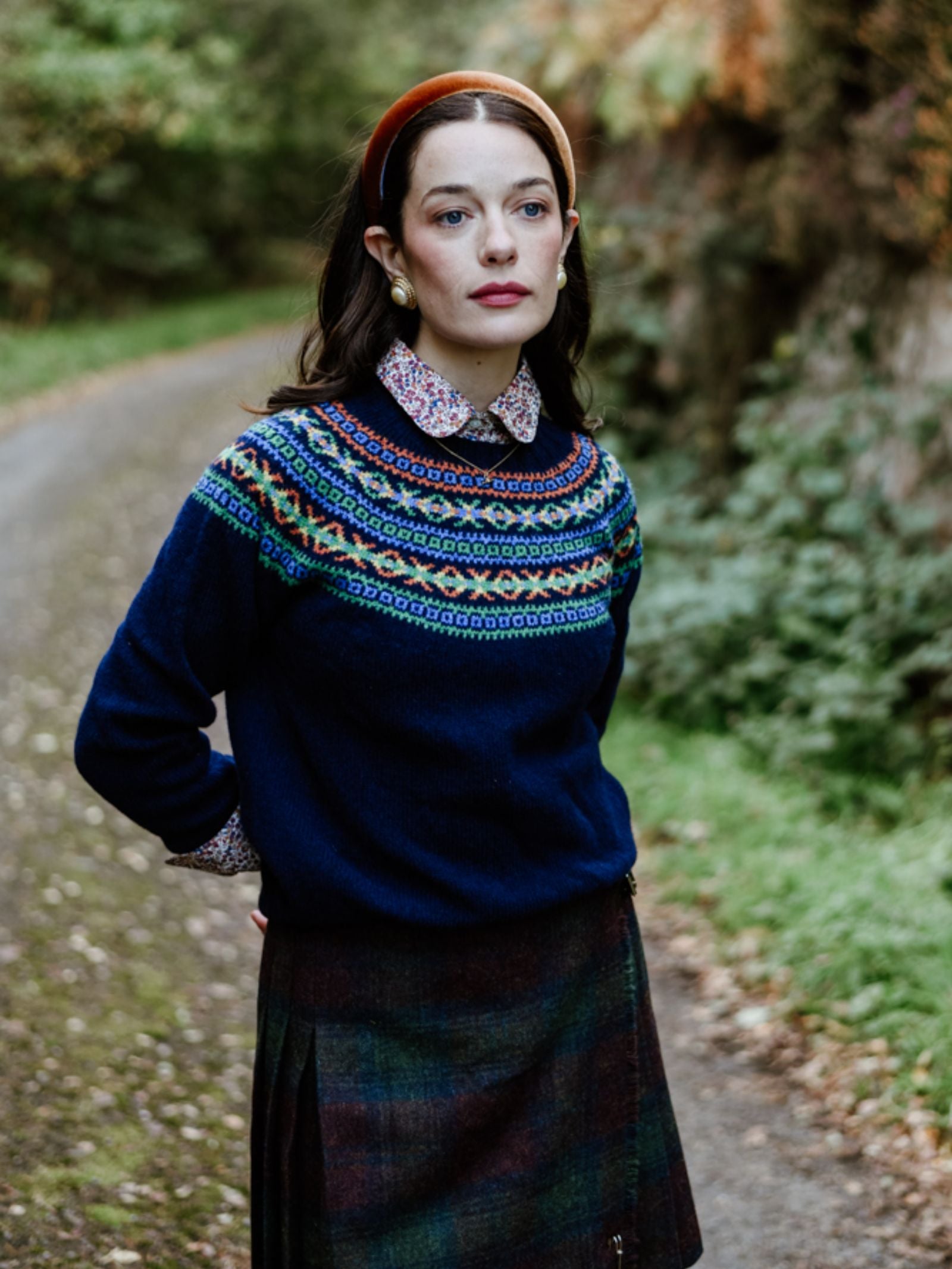 A woman stands on a forest path, thoughtfully gazing into the distance. She wears a Campbell's of Beauly Auld Stock Fairisle Crew Jumper in navy, layered over a collared shirt with a dark plaid skirt and an orange headband.