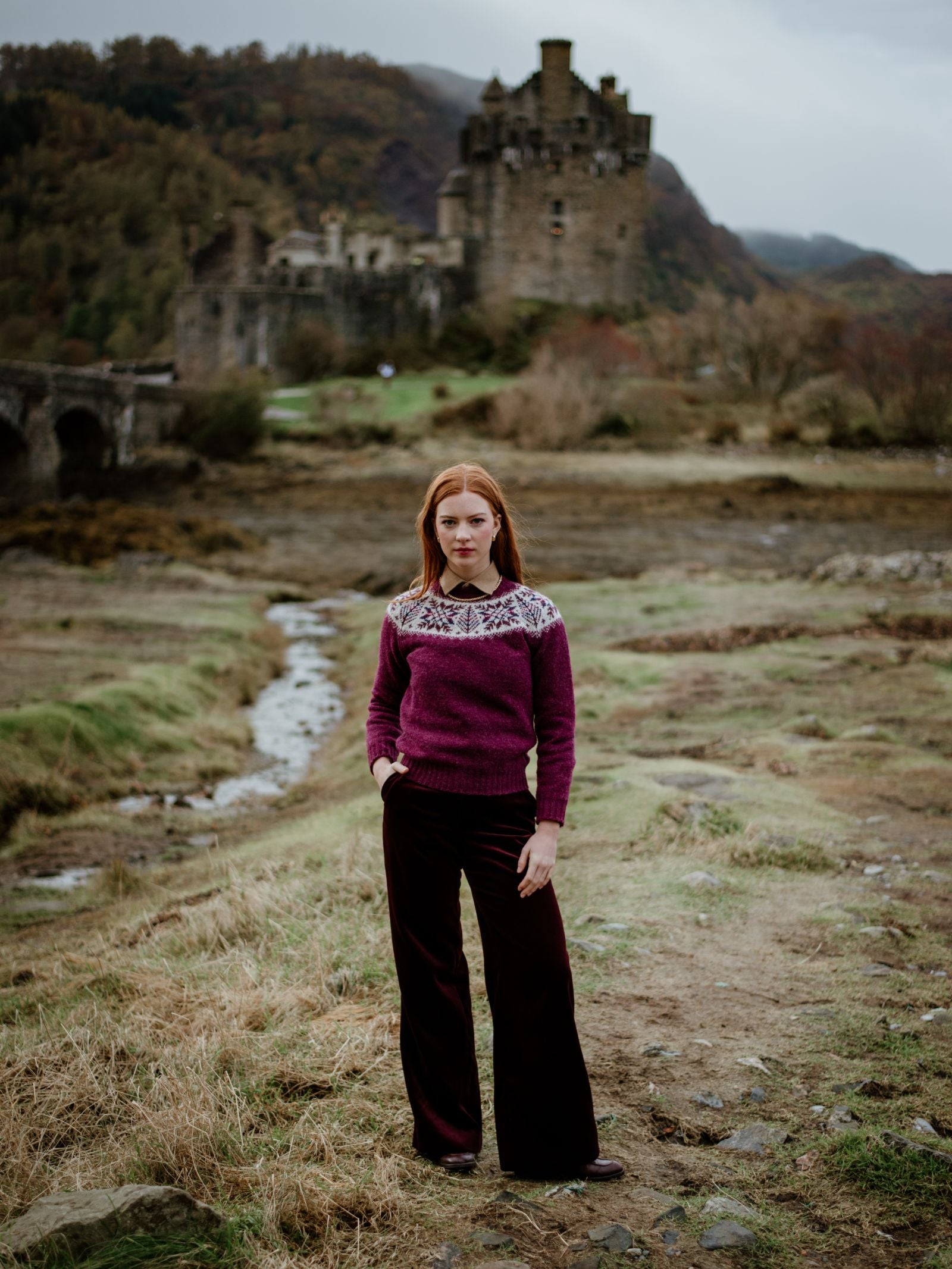A woman with red hair wears the Campbells of Beauly Shetland Wool Snowflake Crew Jumper and wide-legged pants, standing on grassy terrain with hands in pockets; a blurred stone castle is visible in the background.