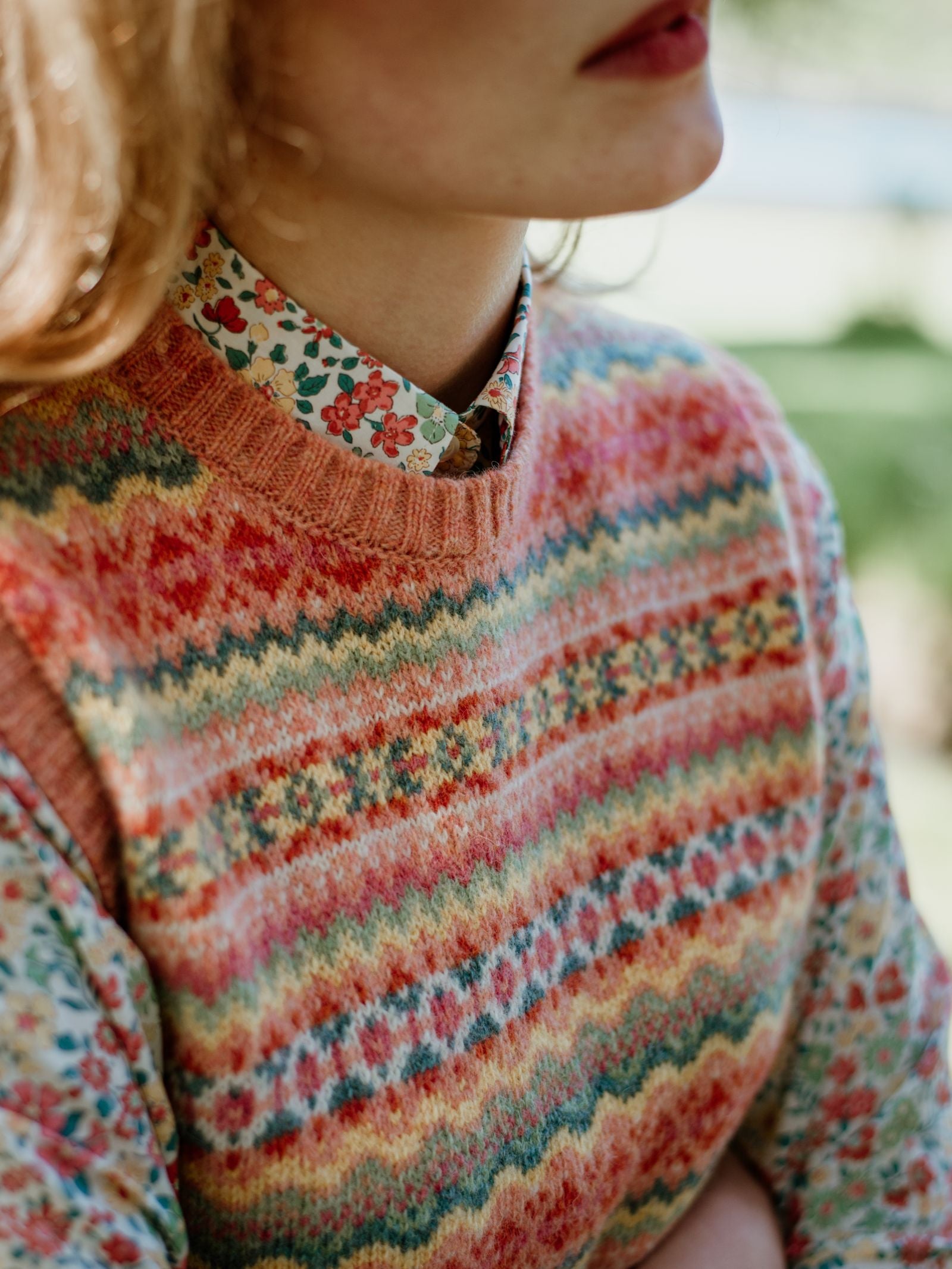 Close-up of a person wearing Campbells of Beauly Fairisle Crew Tank in Shetland wool over a floral button-up shirt. The multicolored traditional pattern stands out, with the face partly visible and blurred greenery in the background.
