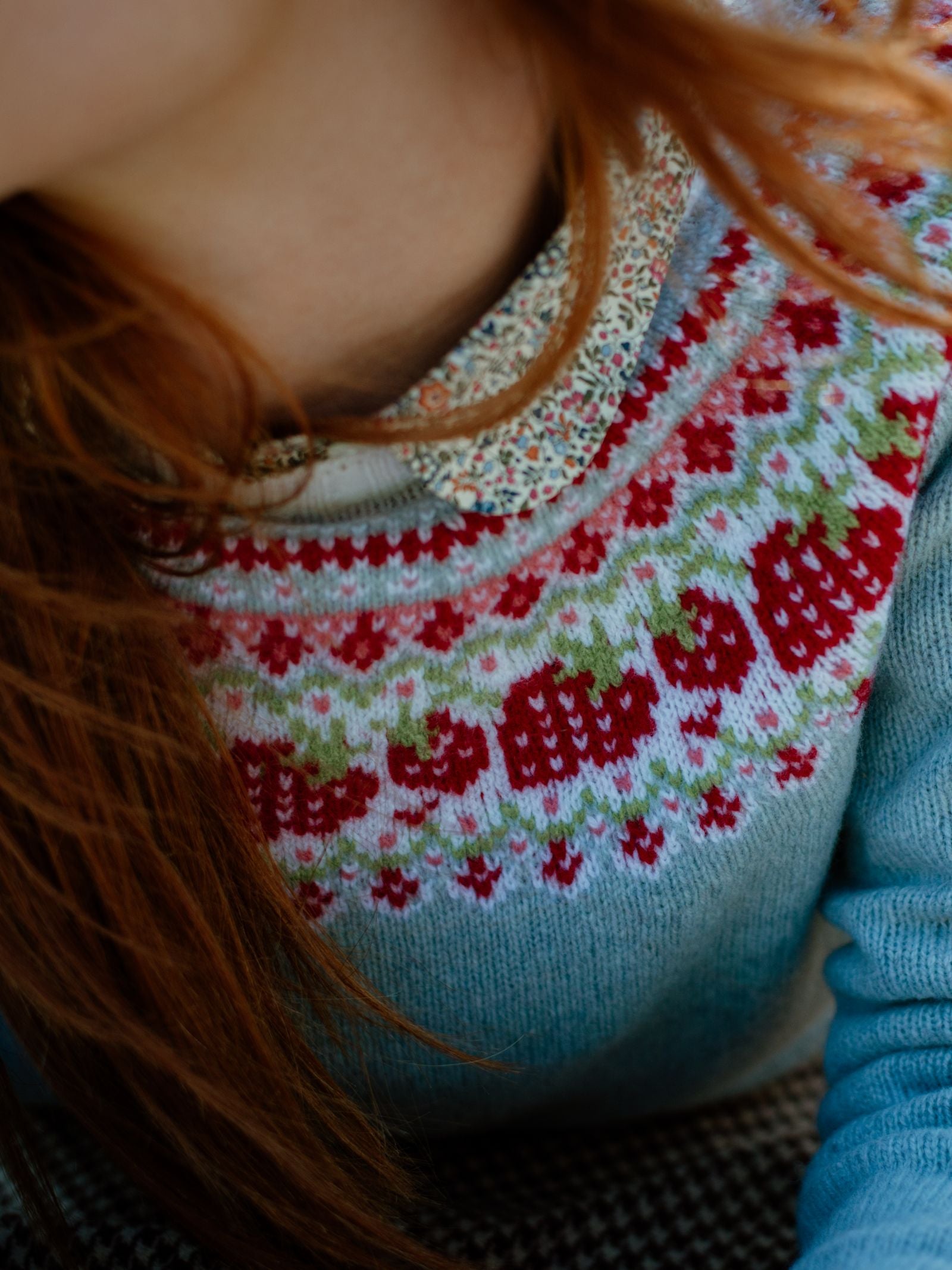 Close-up of a person with long red hair wearing the Campbells of Beauly Geelong Lambswool Strawberry Yoke Jumper in blue, featuring a red and green strawberry pattern over a floral-collared shirt.