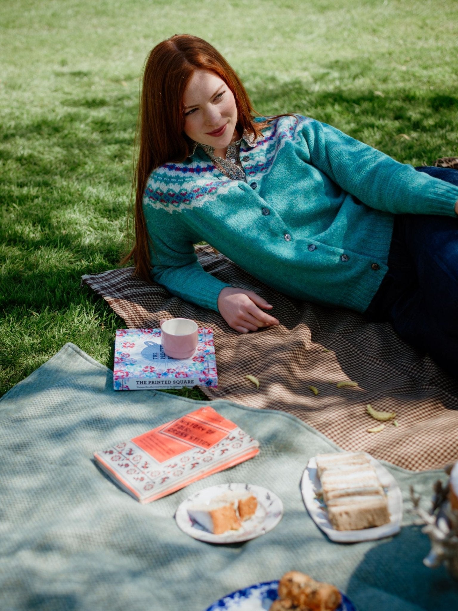 A woman with long red hair lounges on a picnic blanket wearing the Campbell’s of Beauly Shetland Fairisle Cardigan, crafted from Pure New Wool, surrounded by books, snacks, and tea for a relaxed Scottish knitwear-inspired scene.