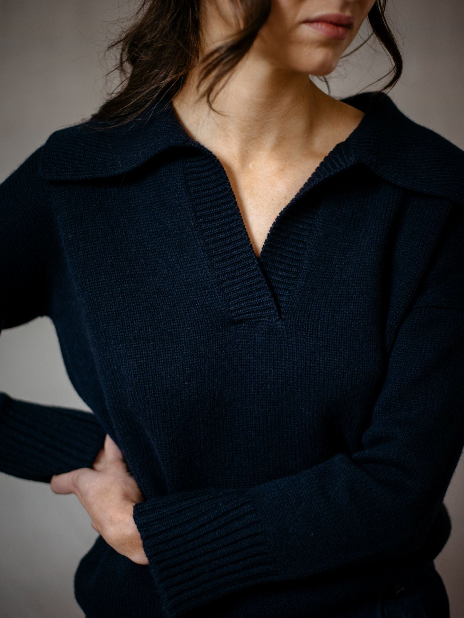 A woman with wavy brown hair, wearing the Campbells of Beauly Cashmere Shirt Jumper in dark navy, stands with her arms crossed and face partly visible. The soft cashmere piece is inspired by the Scottish Borders.