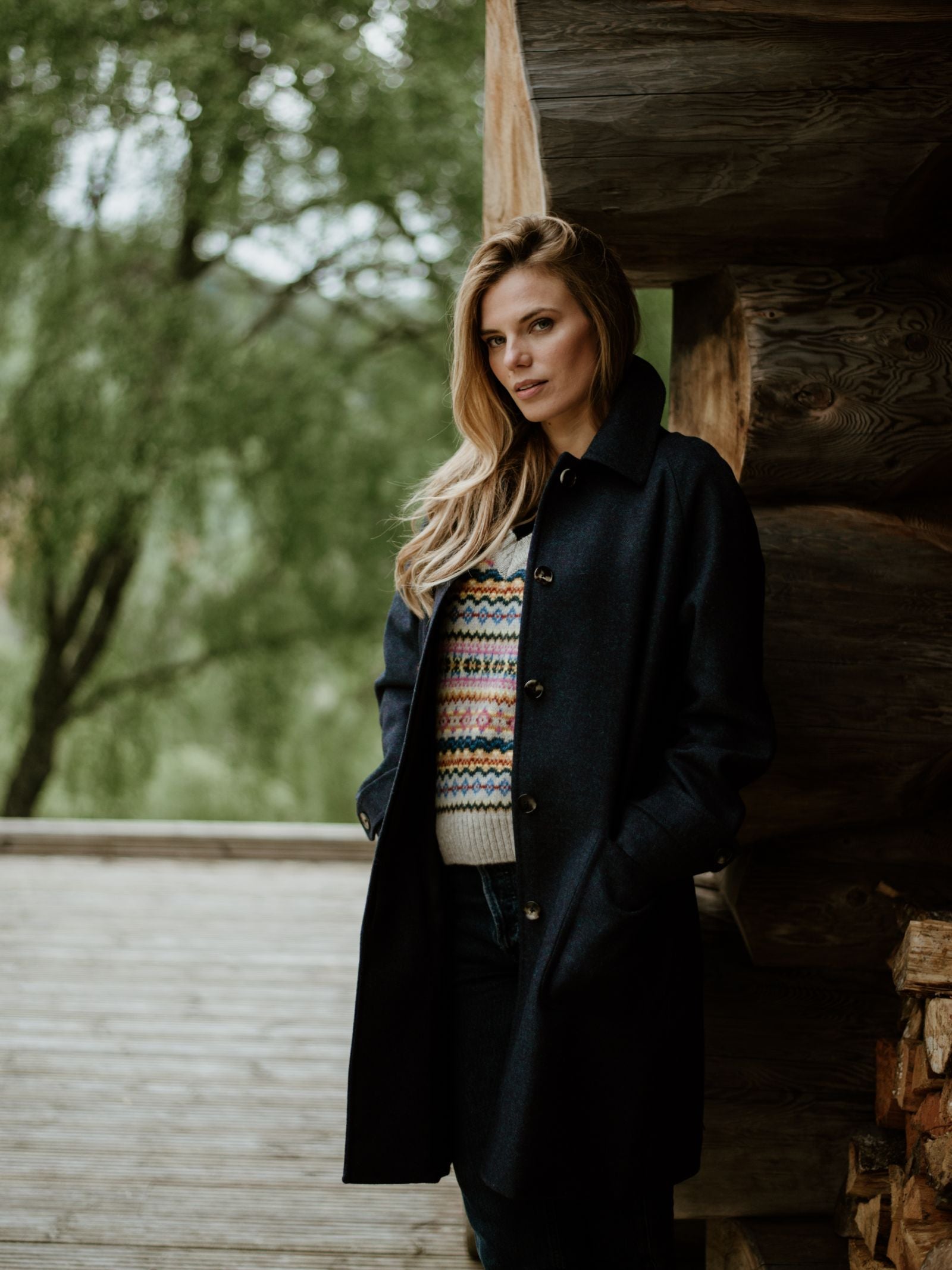 A woman with long blonde hair stands on a wooden porch wearing the Midnight Collar Coat by Campbells of Beauly over a patterned sweater, one hand in her pocket. Trees and greenery appear in the background.