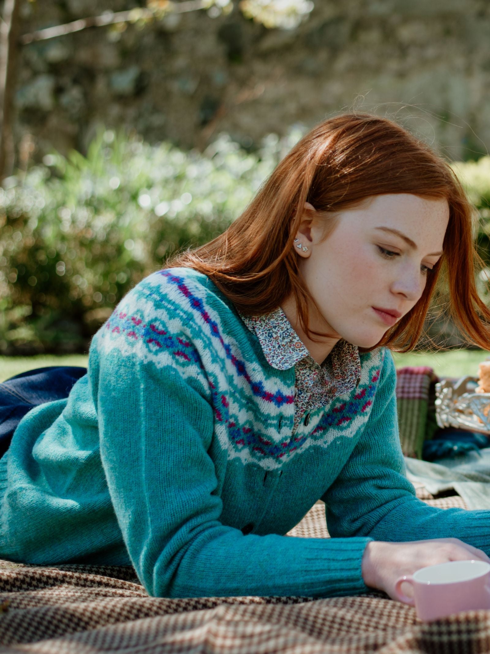 A young woman with long red hair wears a Campbells of Beauly Shetland Fairisle Cardigan while lying on a picnic blanket outdoors, thoughtfully looking down and holding a cup. Sunlight and greenery fill the background.