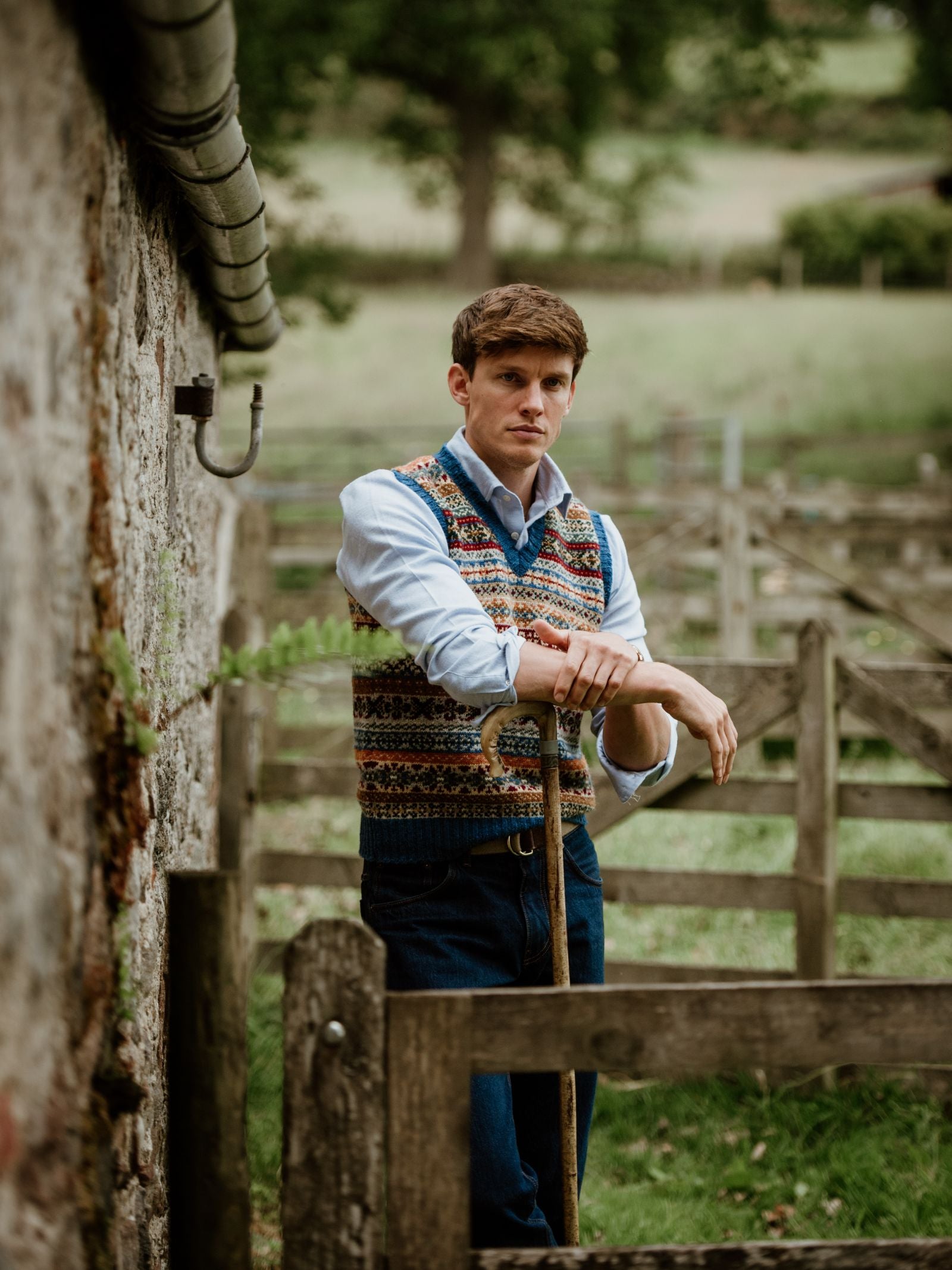 A man in a Campbells of Beauly Fairisle Slipover and blue shirt stands by a stone wall with a shepherds crook, set against wooden fences and green fields.