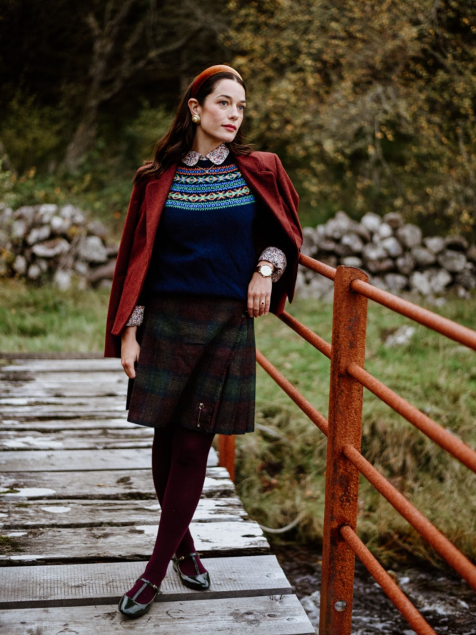 A woman stands on a rustic bridge amid autumn trees, wearing a plaid skirt, burgundy jacket, and the Campbell's of Beauly Fairisle Crew Jumper, expertly knitted in Scotland.