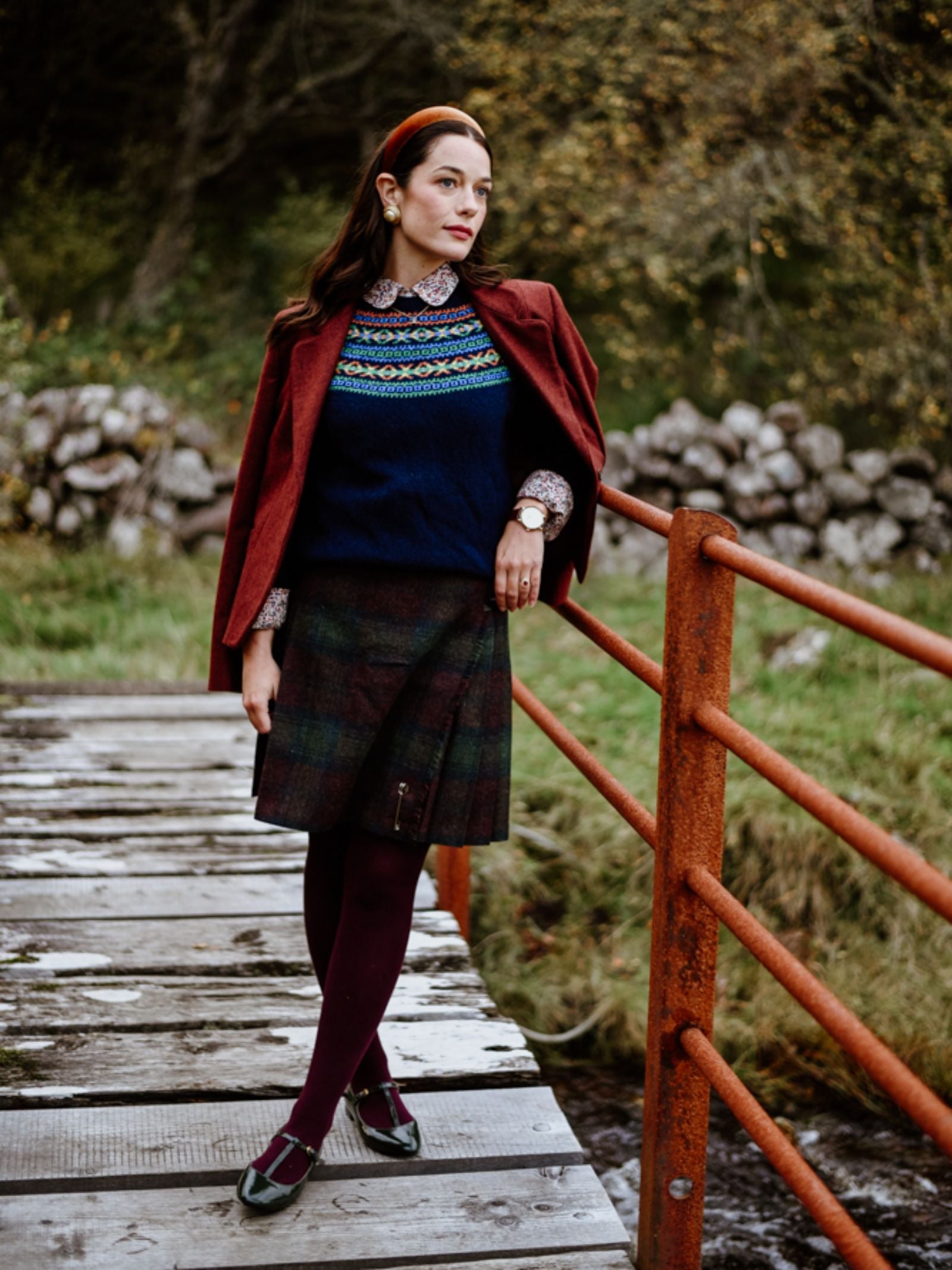 A woman stands on a rustic wooden bridge outdoors, wearing the House Jacket by Campbell's of Beauly. Autumn trees and a stone wall are visible in the background.