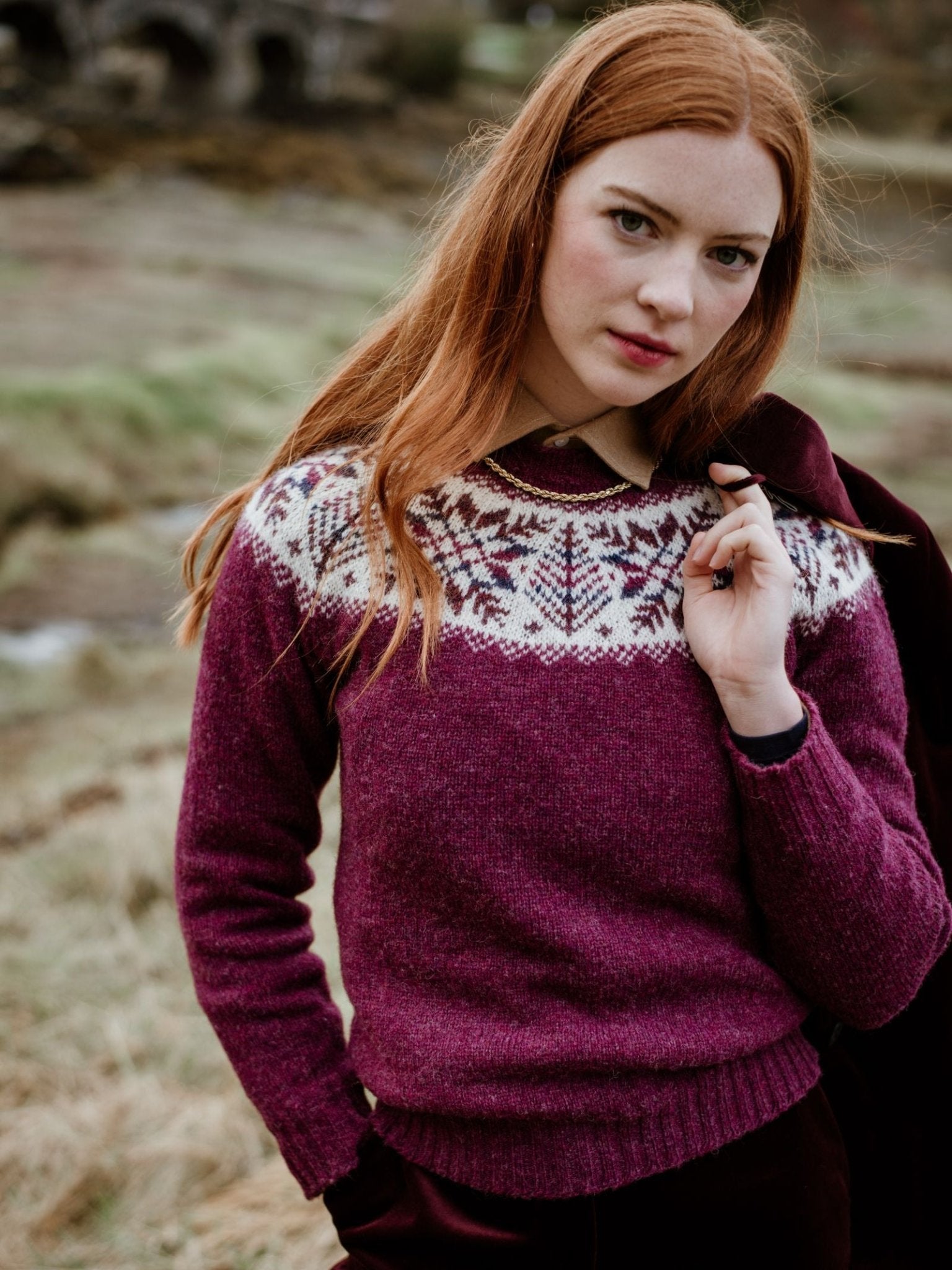 A woman with long red hair stands outdoors on grassy terrain, wearing the Campbells of Beauly Shetland Wool Snowflake Crew Jumper, featuring a maroon base and white snowflake pattern yoke. She holds a coat over her shoulder and looks at the camera.