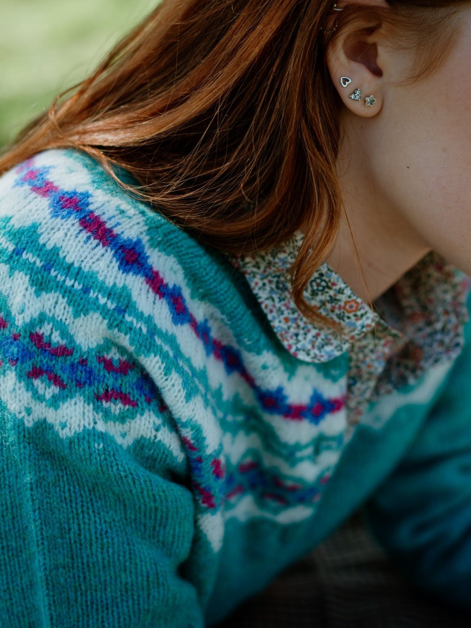 A person with long auburn hair wears the Campbells of Beauly Shetland Fairisle Cardigan over a floral collared shirt, accessorized with multiple small earrings. The image highlights their upper shoulder, neck, and hair.