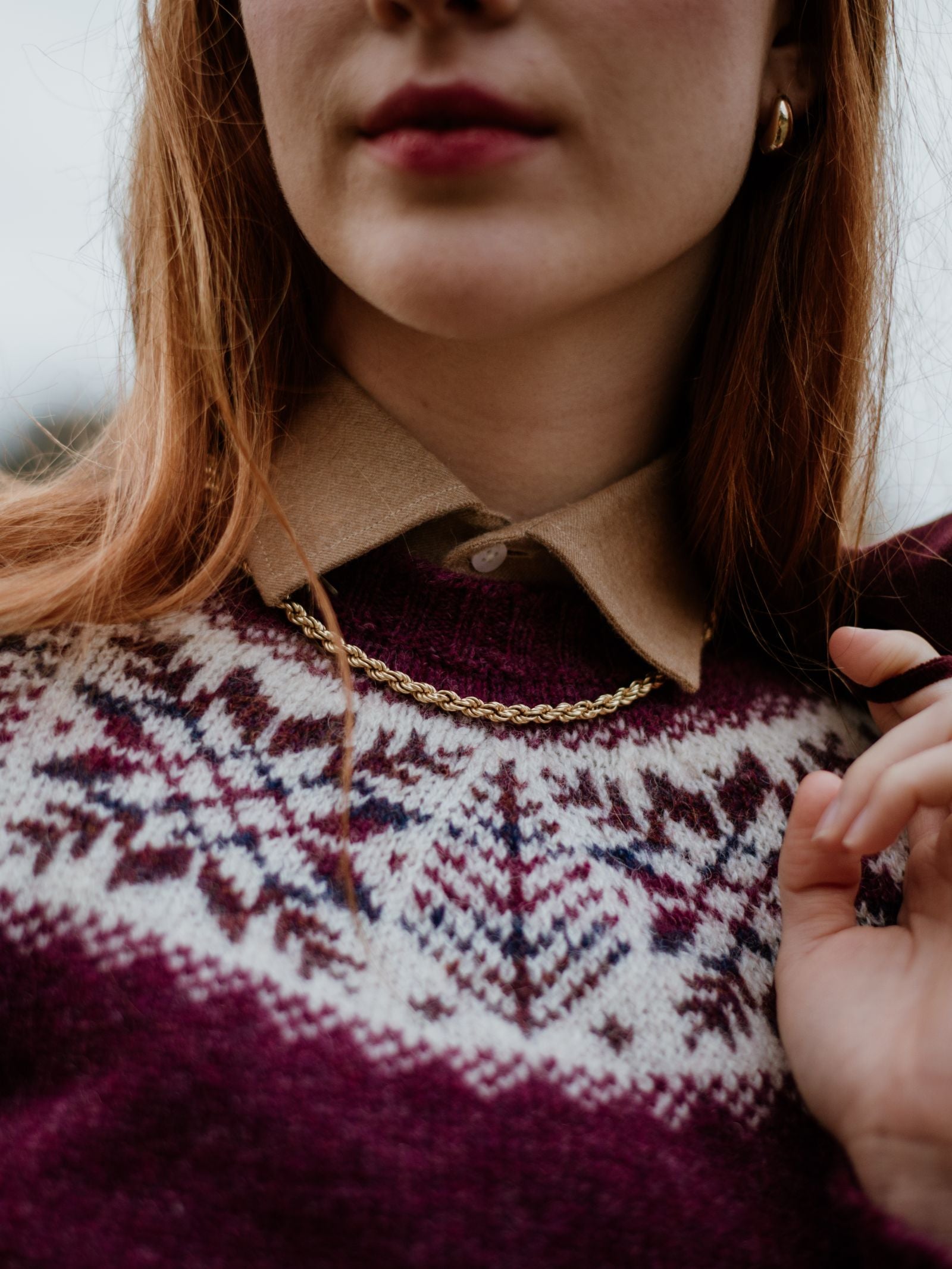 A close-up of a woman wearing the Campbells of Beauly Shetland Wool Snowflake Crew Jumper in maroon and white over a tan collared shirt, styled with a gold chain necklace, small gold hoops, and berry lipstick.