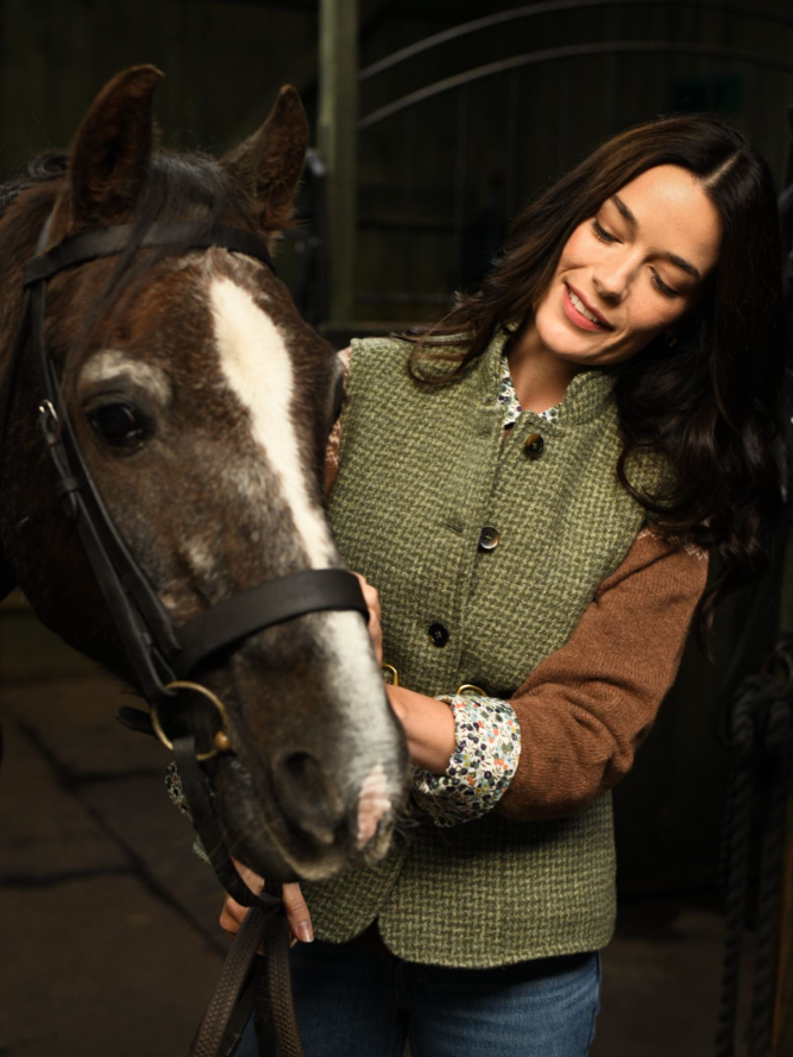 A smiling woman with long dark hair pets a brown horse with a white facial stripe. She wears a Campbell's of Beauly Harris Tweed Houndstooth Gilet and stands in a dimly lit stable.