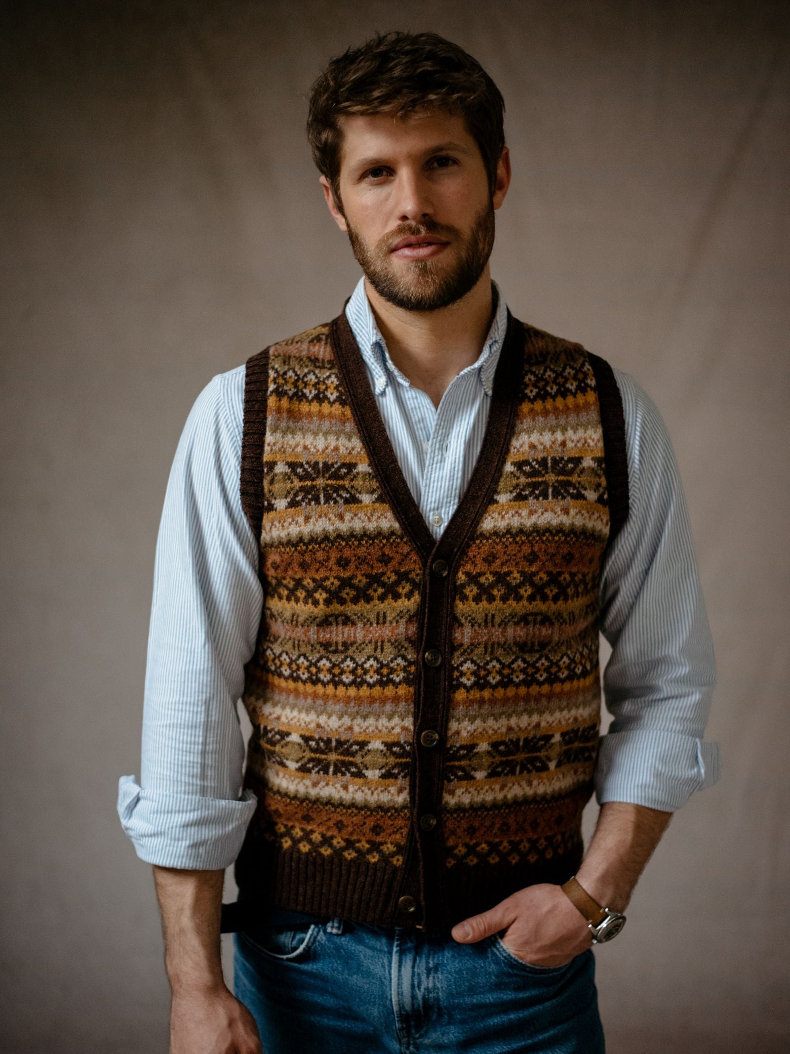 A man with short brown hair and a beard models the Campbells of Beauly Fairisle V-Neck Waistcoat over a striped button-down shirt and jeans, standing against a plain neutral background.