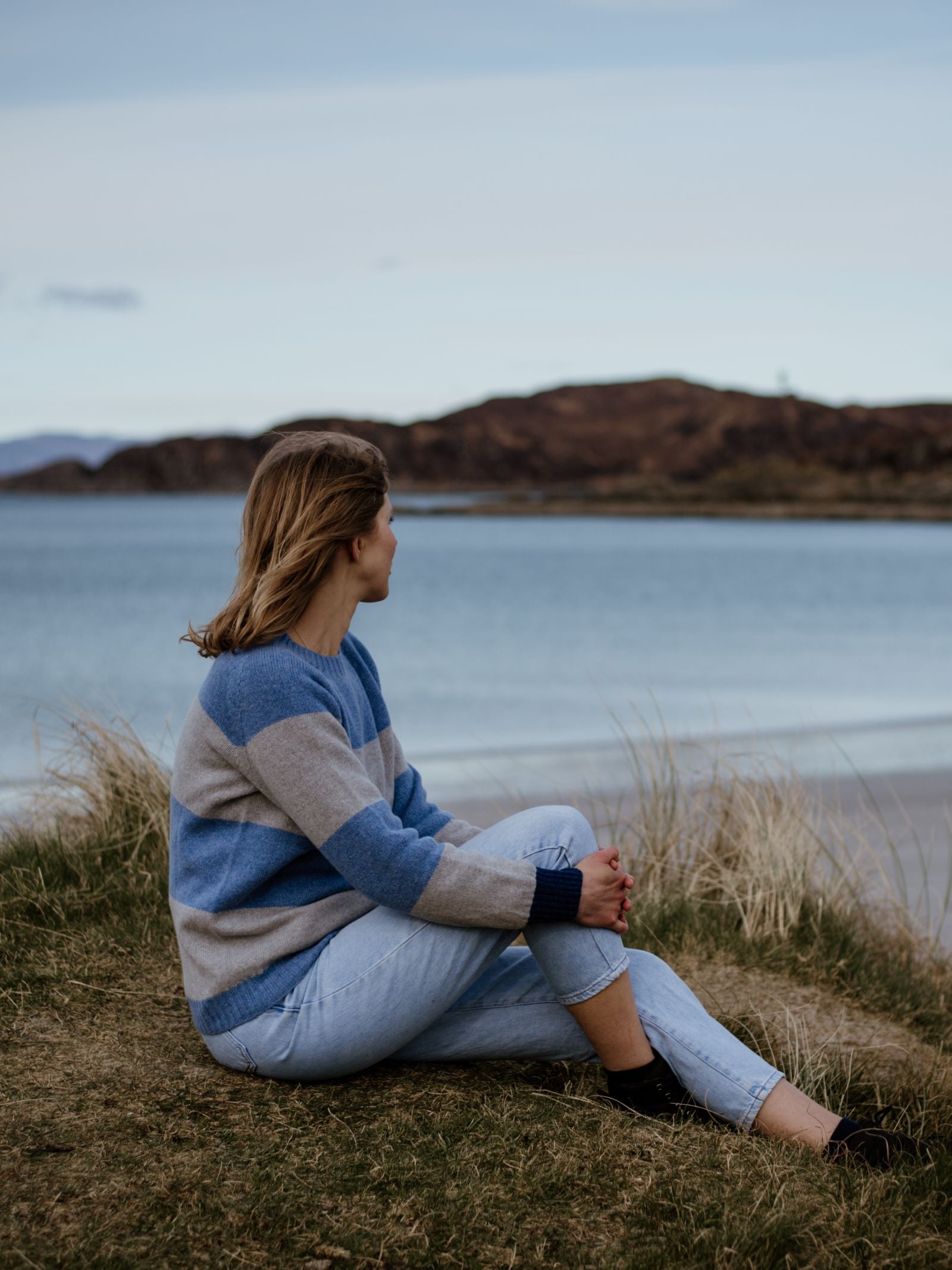A person with light brown hair sits on grassy dunes, wearing the Campbells of Beauly Stripe Crew Jumper—a blue and gray Scottish-knit crew neck—gazing over calm water with hills beyond beneath a cloudy sky.