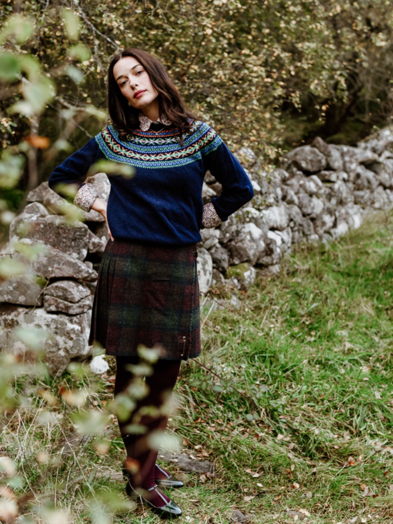 A woman stands by a stone wall amid greenery, wearing Campbell's of Beauly Fairisle Crew Jumper in Geelong lambswool, paired with a plaid skirt and dark tights. She looks relaxed with her hands on her hips.