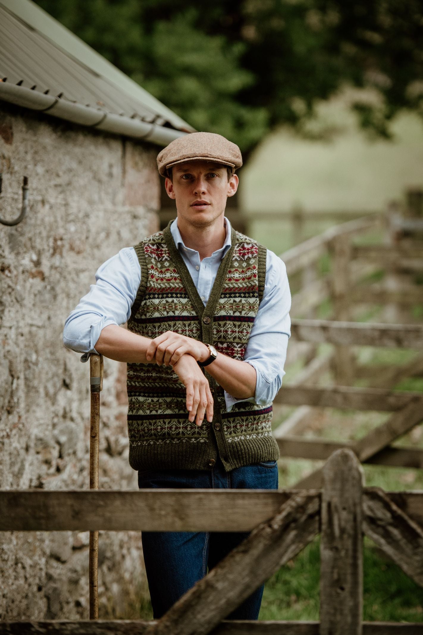A man in a light blue shirt, flat cap, and Campbells of Beauly Fairisle V-Neck Waistcoat stands by a wooden fence holding a cane, with a stone wall and green trees behind him.