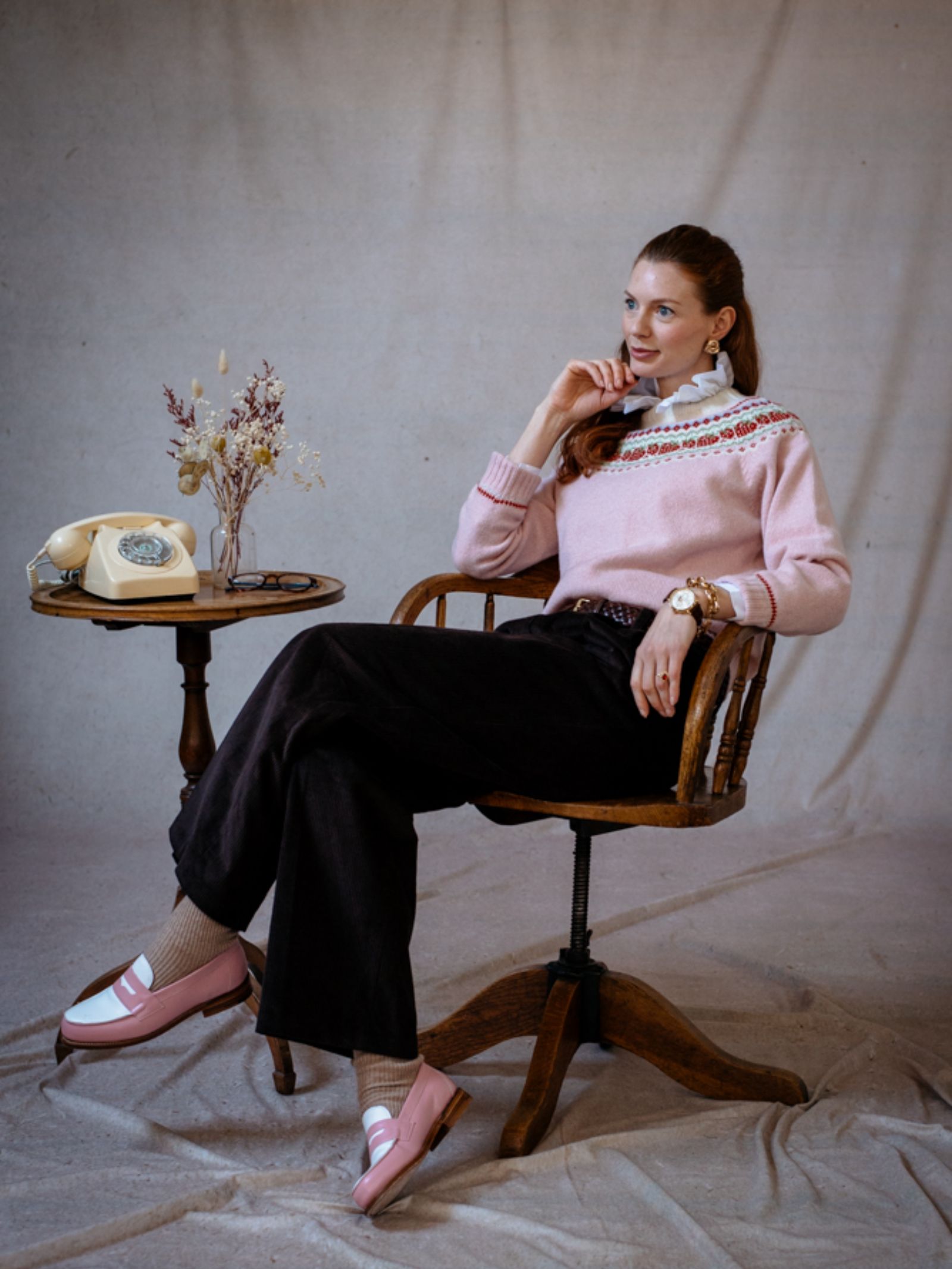 A woman in a Campbells of Beauly Geelong Lambswool Strawberry Yoke Jumper sits on a wooden chair by a table with a vintage rotary phone and dried flowers, posing with one hand on her chin and wearing wide-leg pants and pink shoes.