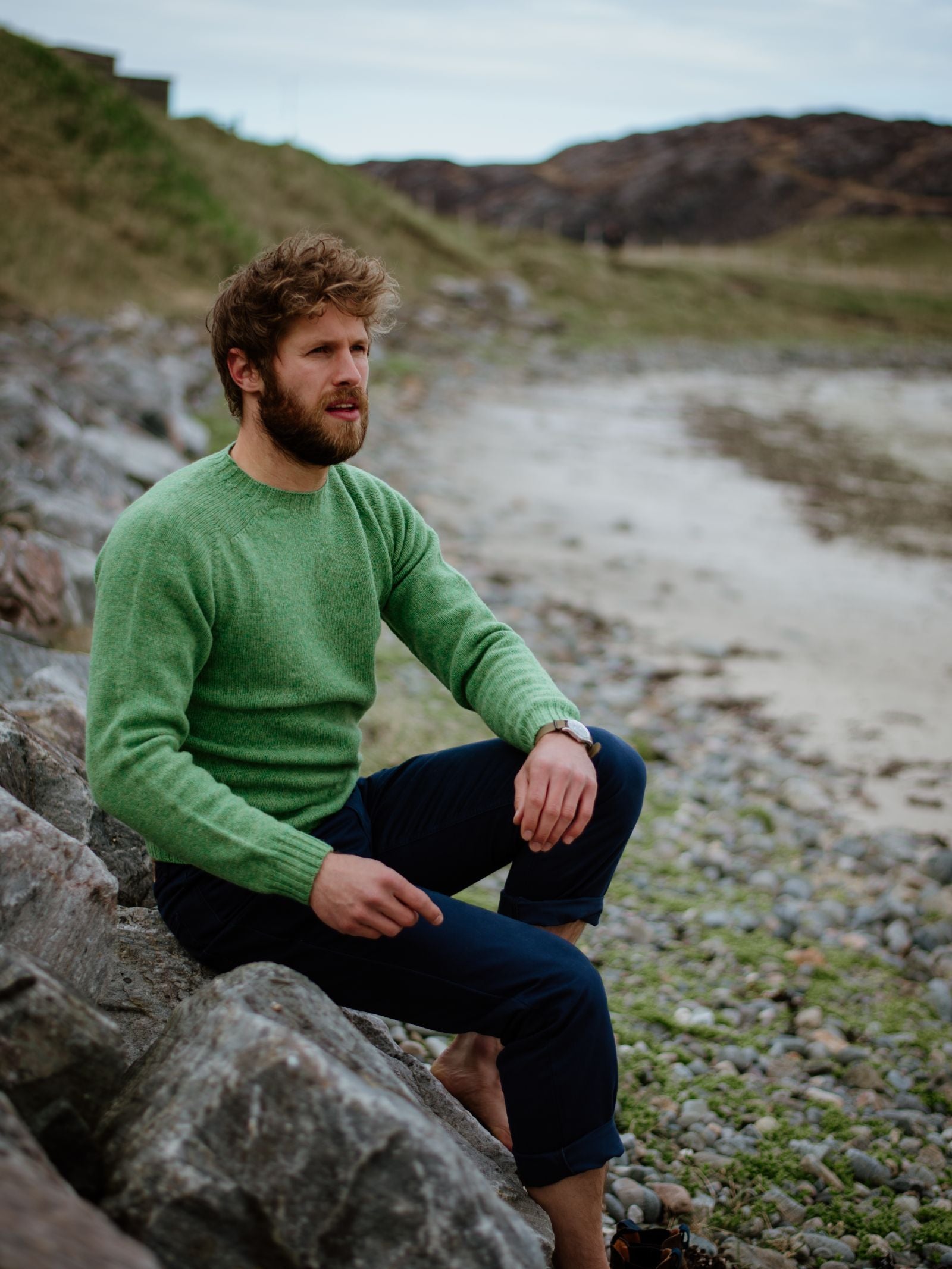 A bearded man wearing a Campbells of Beauly Shetland Jumper and dark pants sits barefoot on rocky ground by the beach, gazing thoughtfully. Grassy hills and a cloudy sky provide the scenic background.
