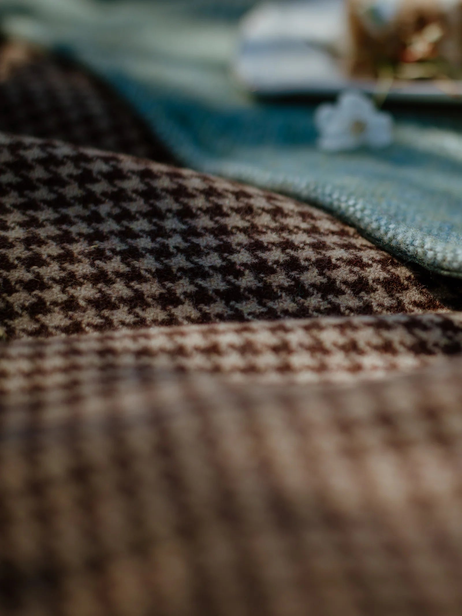 A close-up of the Crofters Blanket by Campbells of Beauly shows its brown and beige houndstooth pattern, evoking a classic country style. In the background, a blue-green fabric and blurred white flowers are softly visible.