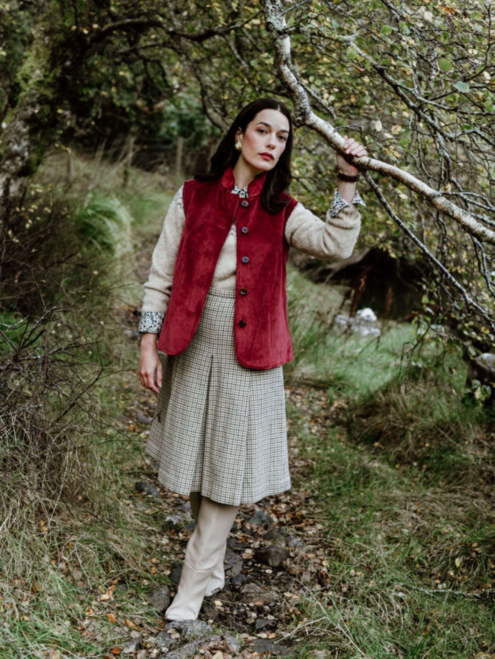 A woman stands on a forest path, wearing the Campbell's of Beauly Corduroy Gilet over a beige blouse and checked skirt with cream boots, exuding country elegance among mossy trees and lush greenery.