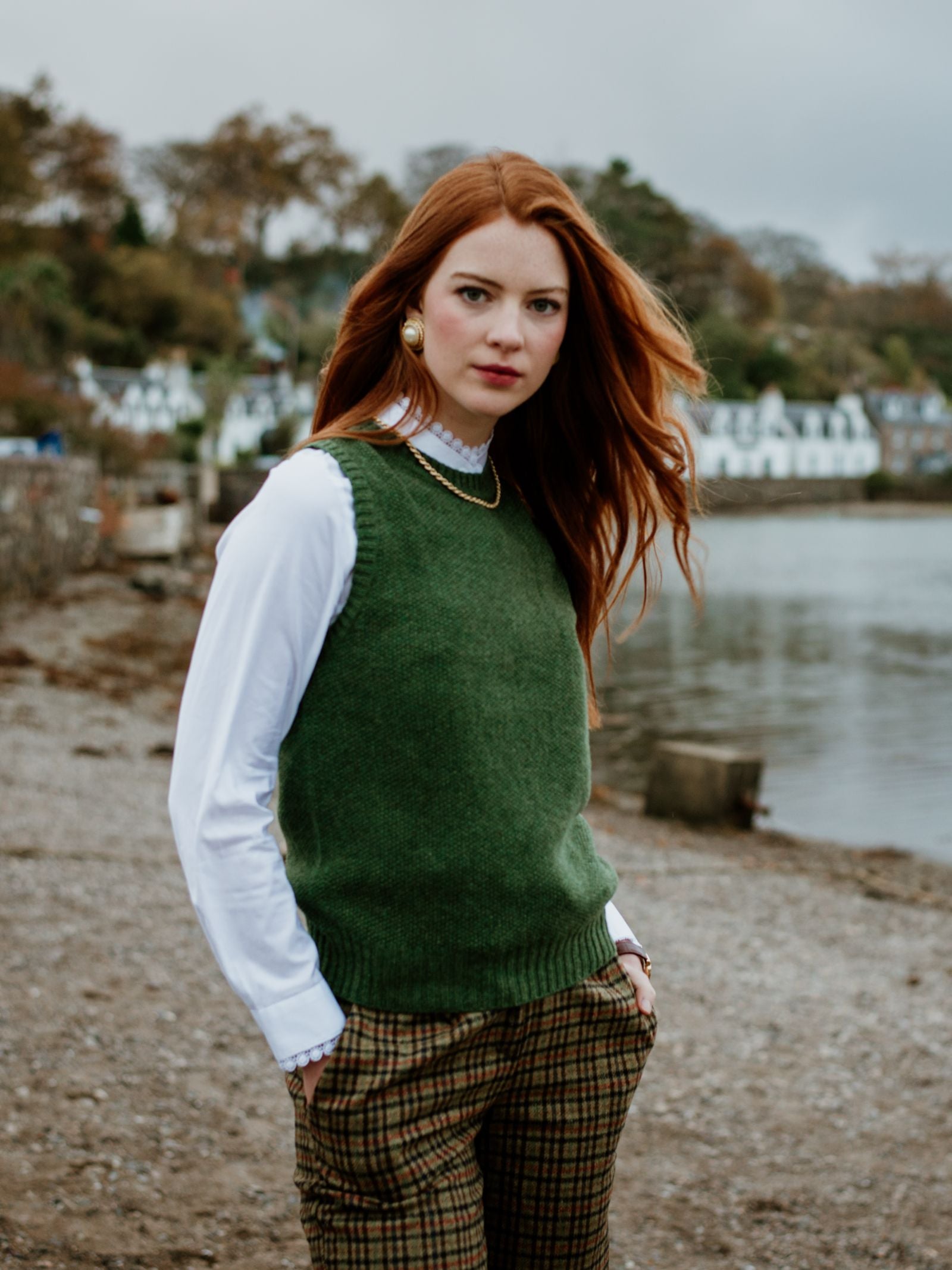 A woman with long red hair wears the Moss Stitch Crew Tank by Campbells of Beauly in green Scottish yarn over a white shirt and plaid pants, standing lakeside with trees and white houses behind her, showing versatile layering.