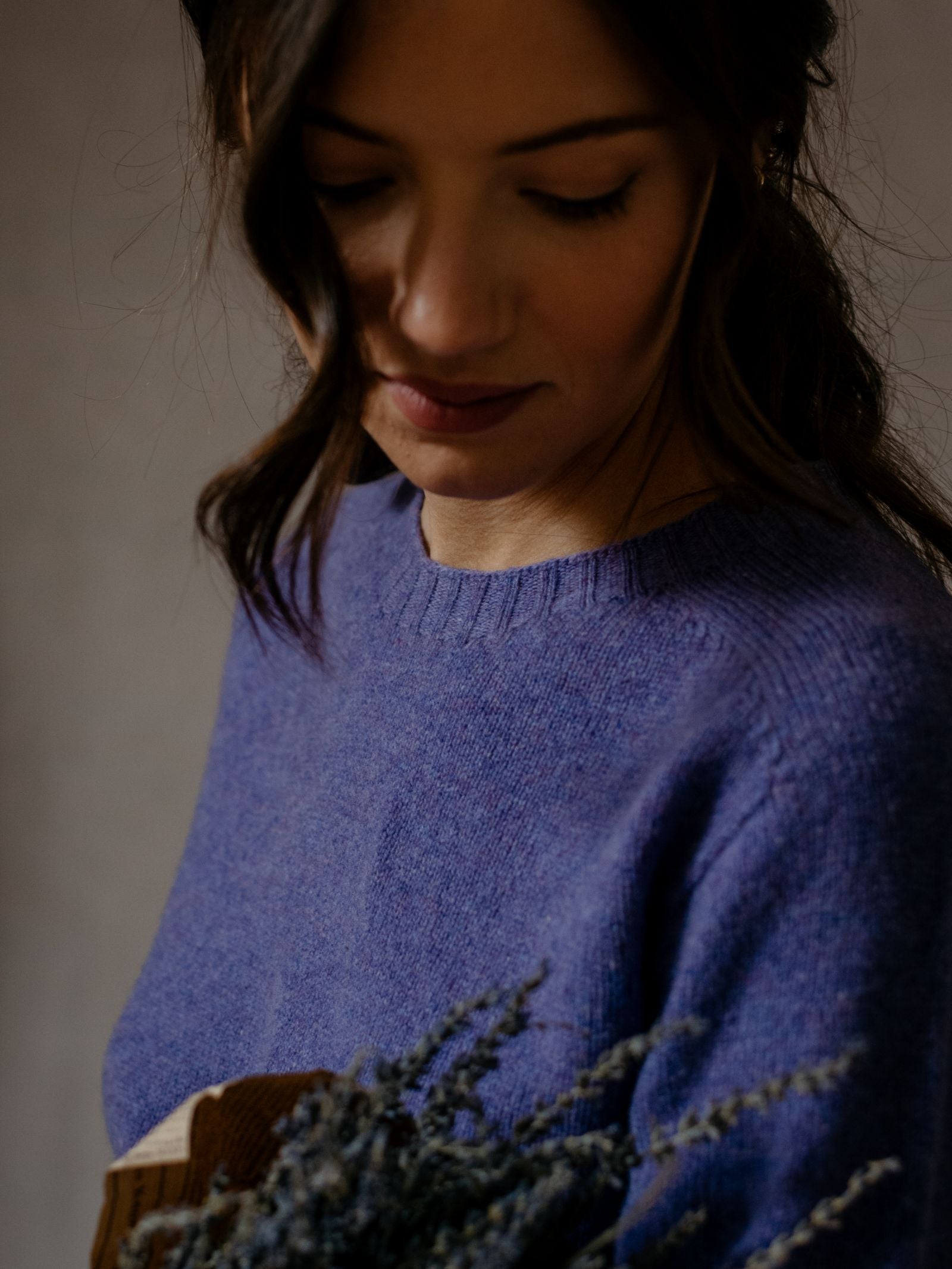 A woman with wavy brown hair holds dried lavender and looks down, wearing a soft purple Geelong Lambswool Crew Neck Jumper by Campbells of Beauly against a neutral background.