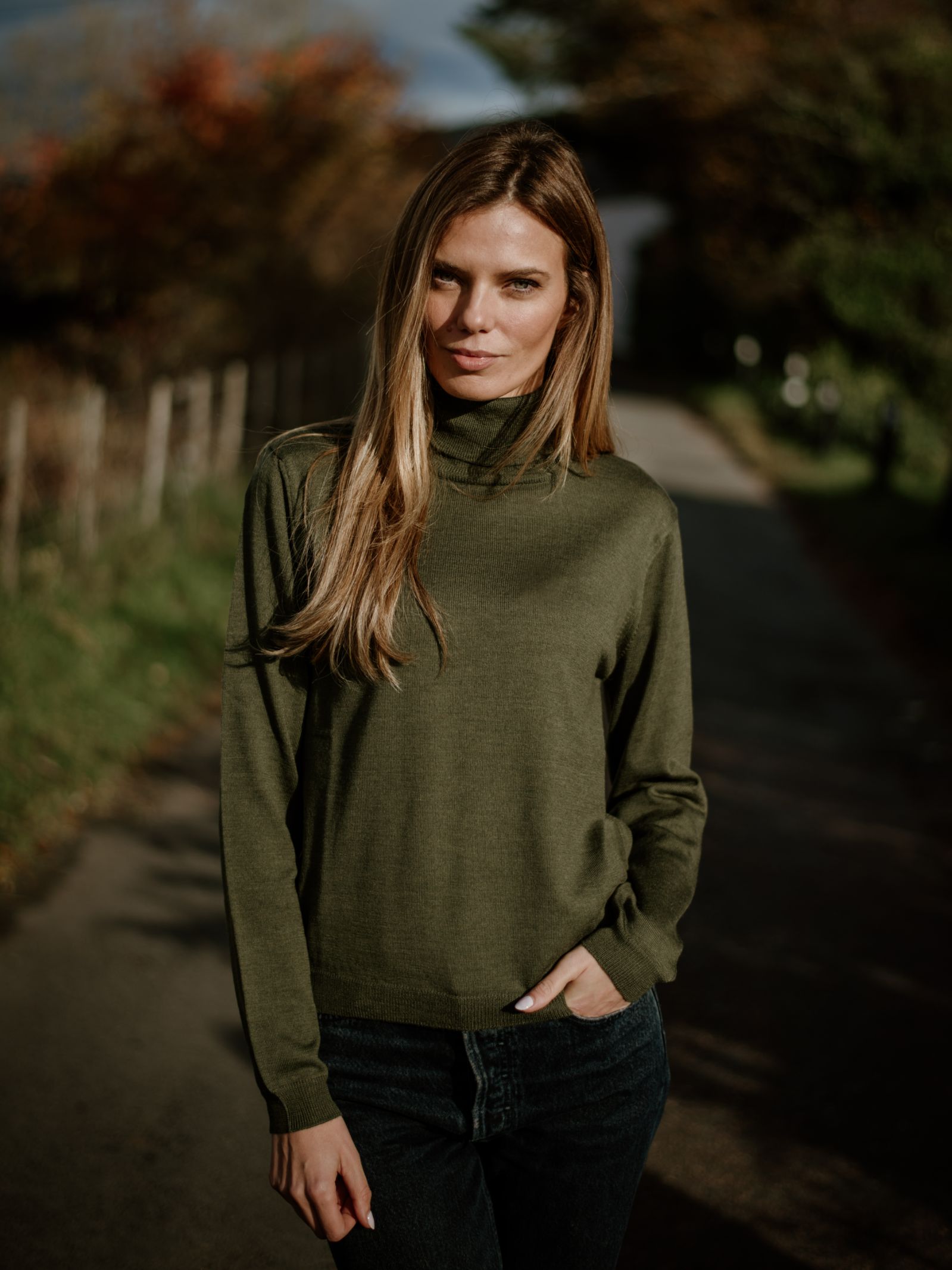 A woman with long blonde hair stands on a shaded path, wearing the Campbells of Beauly Merino Poloneck sweater in green and dark jeans. Autumn trees in the background make this piece perfect for smart seasonal layering.