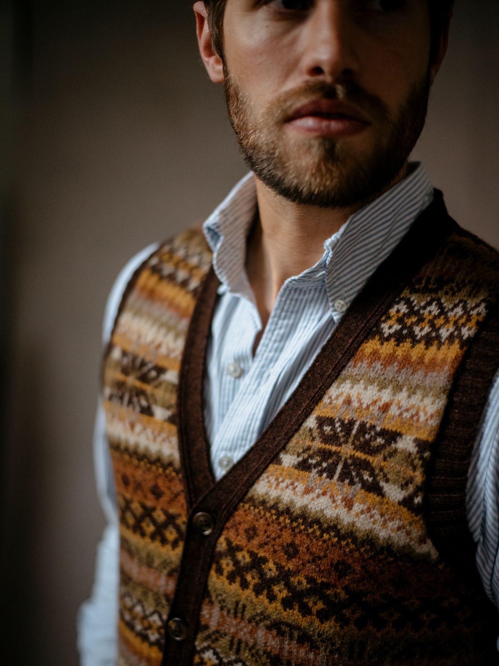 A man with a short beard wears a blue striped shirt beneath a Campbells of Beauly Fairisle V-Neck Waistcoat with geometric patterns. He gazes slightly to the side in gentle, natural light.