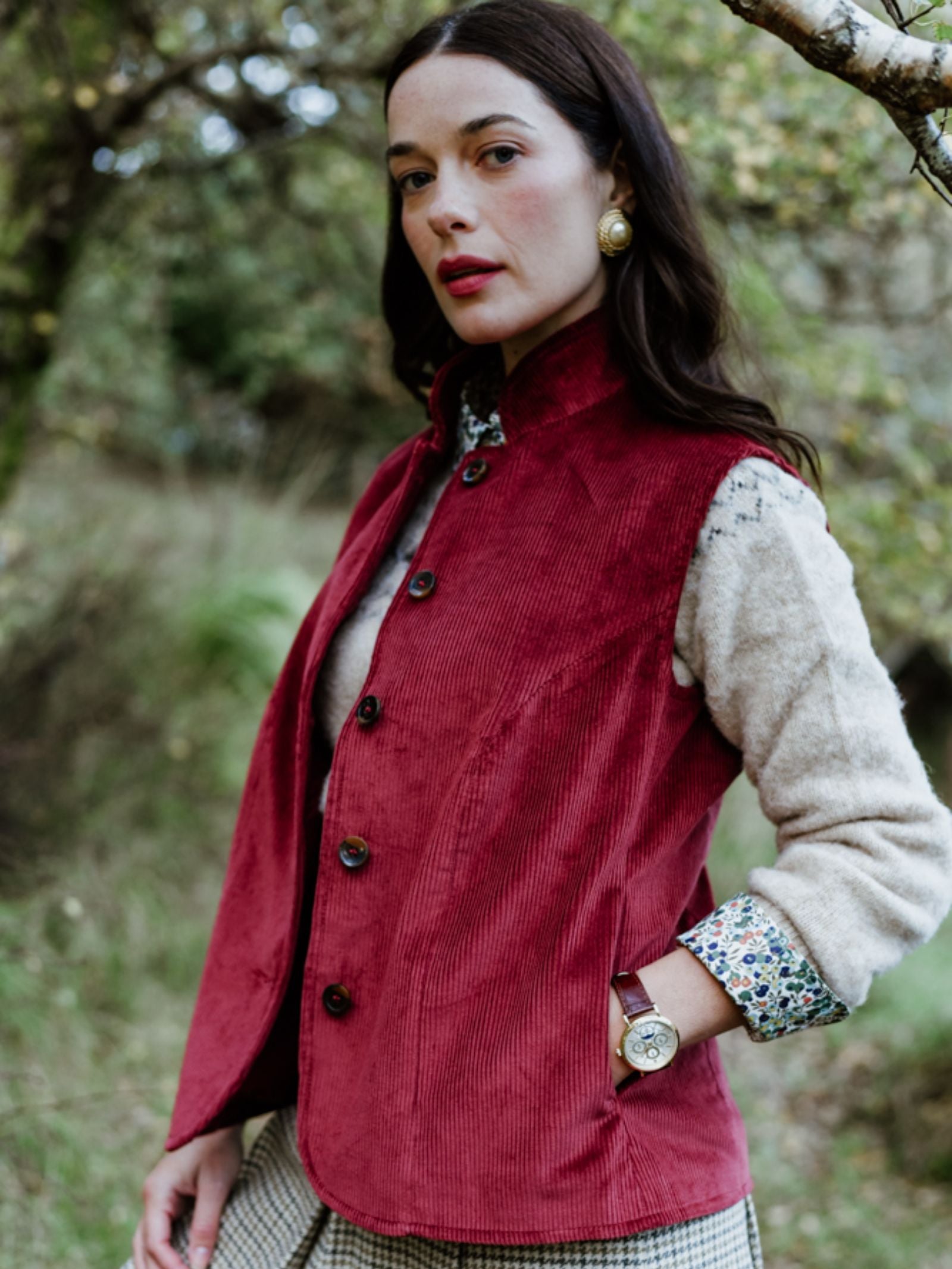 A woman stands outdoors wearing Campbell's of Beauly Corduroy Gilet in red over a beige sweater. With long dark hair, gold earrings, and a wristwatch, she exudes country elegance as she gazes calmly at the camera.